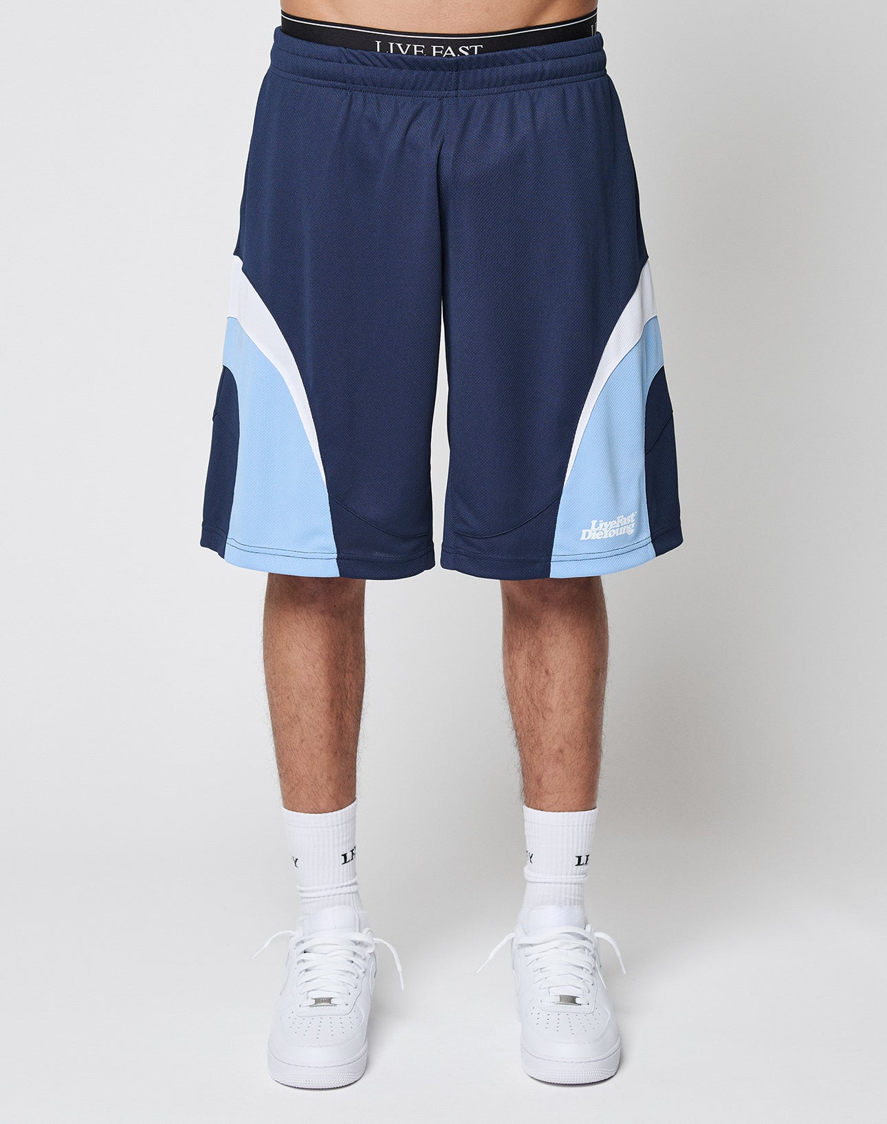 A person models the LIVE FAST DIE YOUNG Basic Trackshorts—navy blue with light blue and white curved accents and an elastic waistband—paired with white socks and sneakers, standing against a plain white background (lower half only visible).