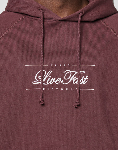 Close-up of a person wearing the LFDY Embroidery Hooded by LIVE FAST DIE YOUNG. This maroon regular fit hoodie features white embroidered text, drawstrings, and is crafted from 100% Baumwolle for comfort.