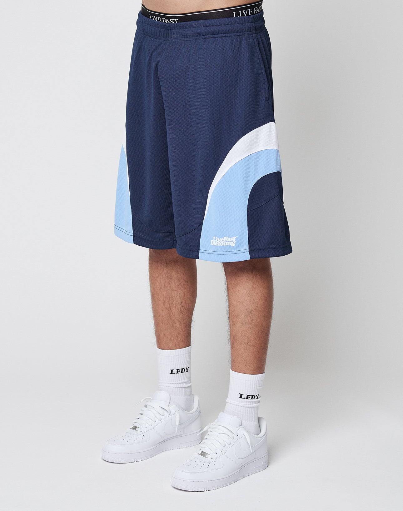 A person wearing LIVE FAST DIE YOUNG Basic Trackshorts in navy and light blue with elastic waistband, paired with white socks featuring LEDY text and white Nike sneakers, is shown from the waist down against a plain white background.