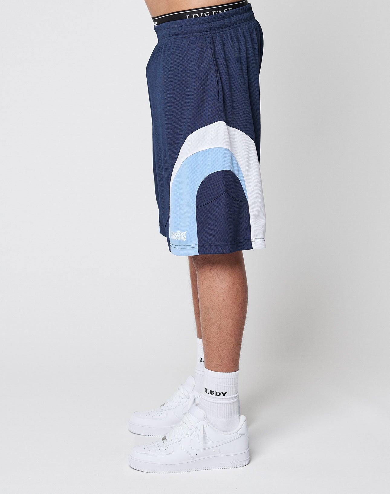 A person stands sideways wearing LIVE FAST DIE YOUNG Basic Trackshorts in navy blue with an elastic waistband, white and light blue curved panels, white socks displaying LEDY text, and white Nike sneakers against a plain white background.
