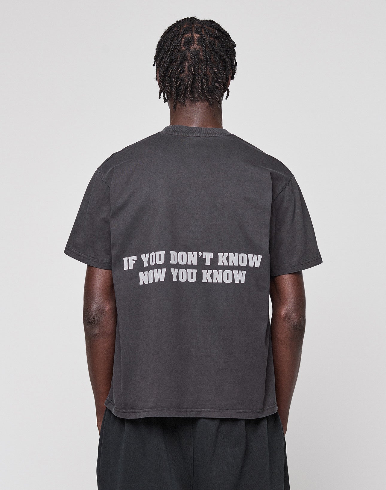 A person with short, twisted hair is seen from the back wearing the LIVE FAST DIE YOUNG Abus Magomedov Tee—a black regular fit t-shirt with "IF YOU DONT KNOW NOW YOU KNOW" in white as a backprint. The background is plain and light-colored.