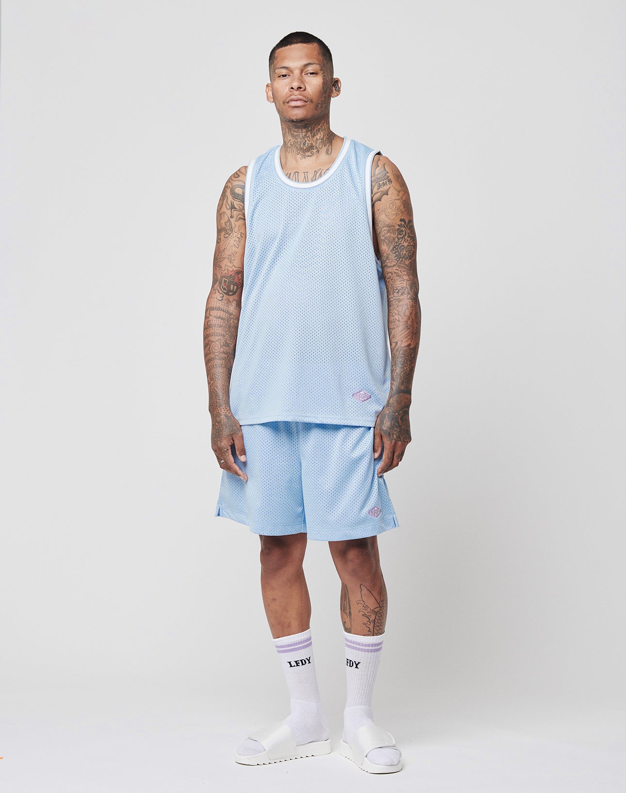 A tattooed man faces forward, wearing the LIVE FAST DIE YOUNG Basic Mesh Tank Top in light blue with matching shorts, white socks featuring black text, and white slide sandals against a plain light background.