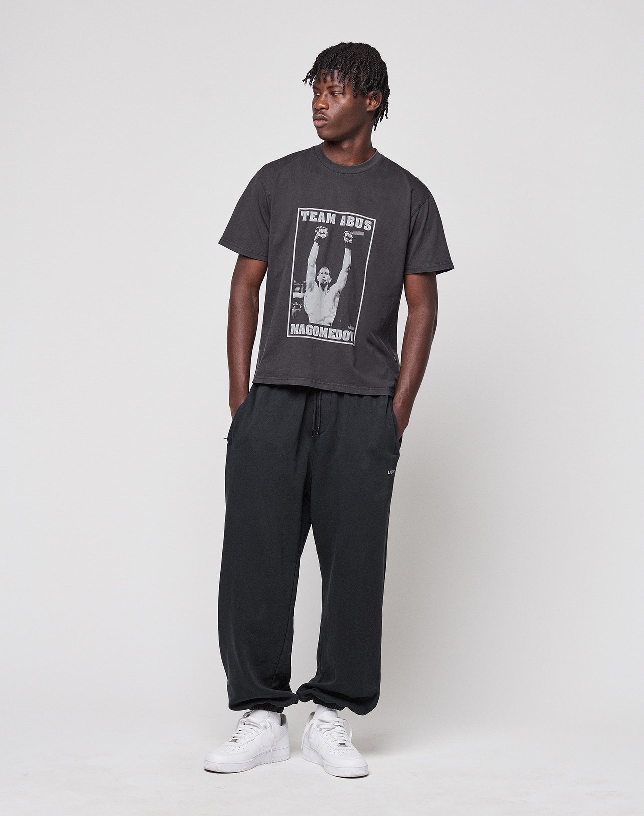 A person with short, twisted hair stands against a plain background, wearing the Abus Magomedov Tee by LIVE FAST DIE YOUNG—a Regular Fit black T-shirt made of 100% Baumwolle with photo and text prints on front and back—plus black sweatpants and white sneakers.