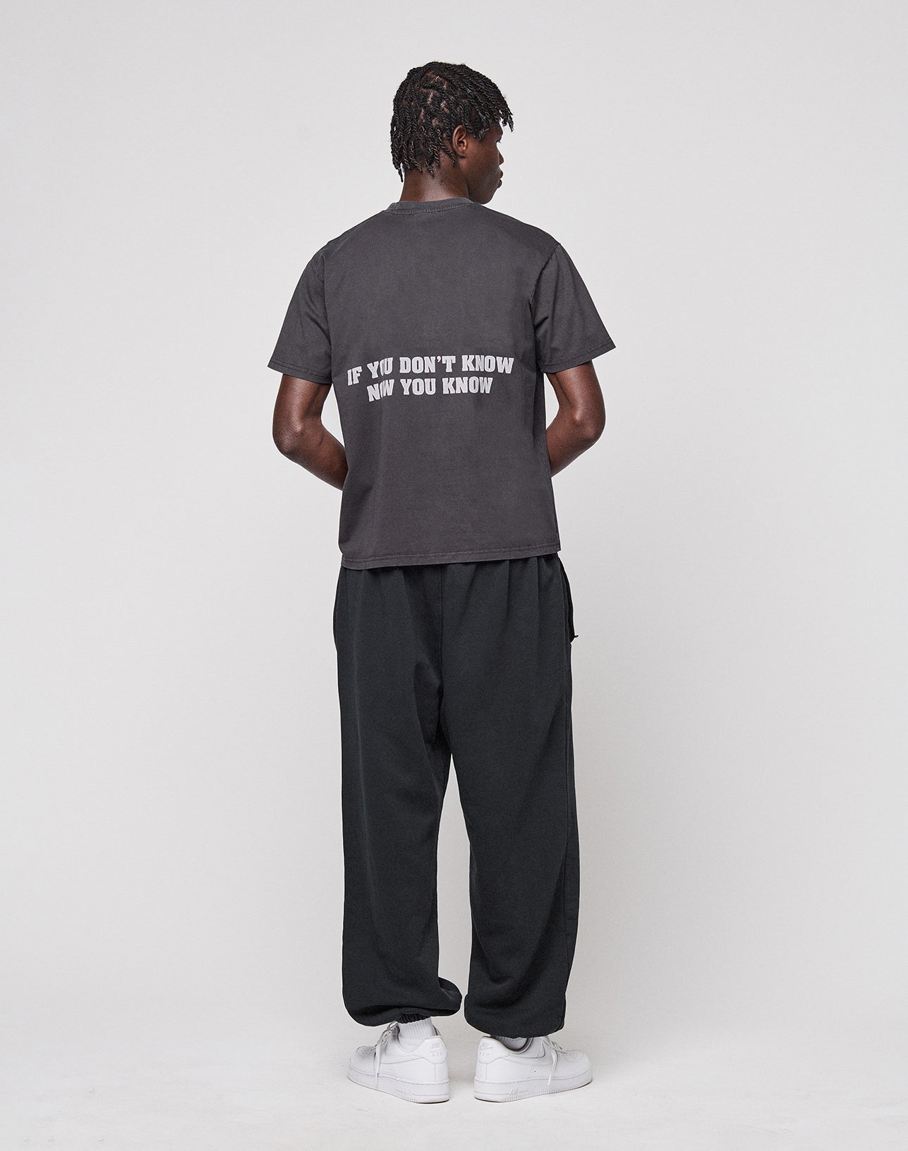 A person faces away, wearing the LIVE FAST DIE YOUNG Abus Magomedov Tee made of 100% Baumwolle with "IF YOU DONT KNOW NOW YOU KNOW" on the back, paired with loose dark pants and white sneakers against a light background.