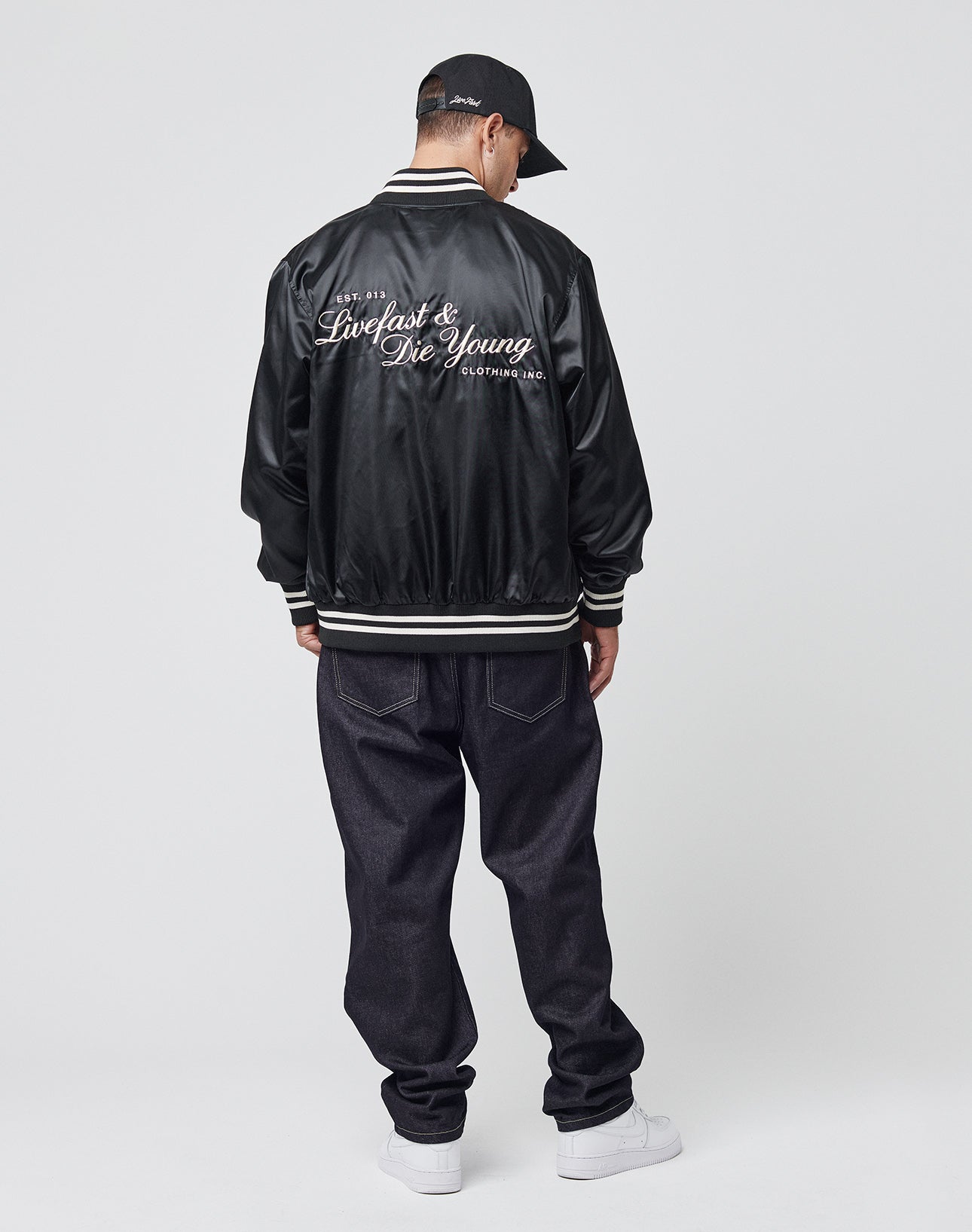 A person faces away, wearing the LIVE FAST DIE YOUNG Satin College Jacket with white trim and Druckknöpfe, paired with dark jeans, white sneakers, and a black cap. Live fast & Die Young is scripted on the back. The background is plain white.