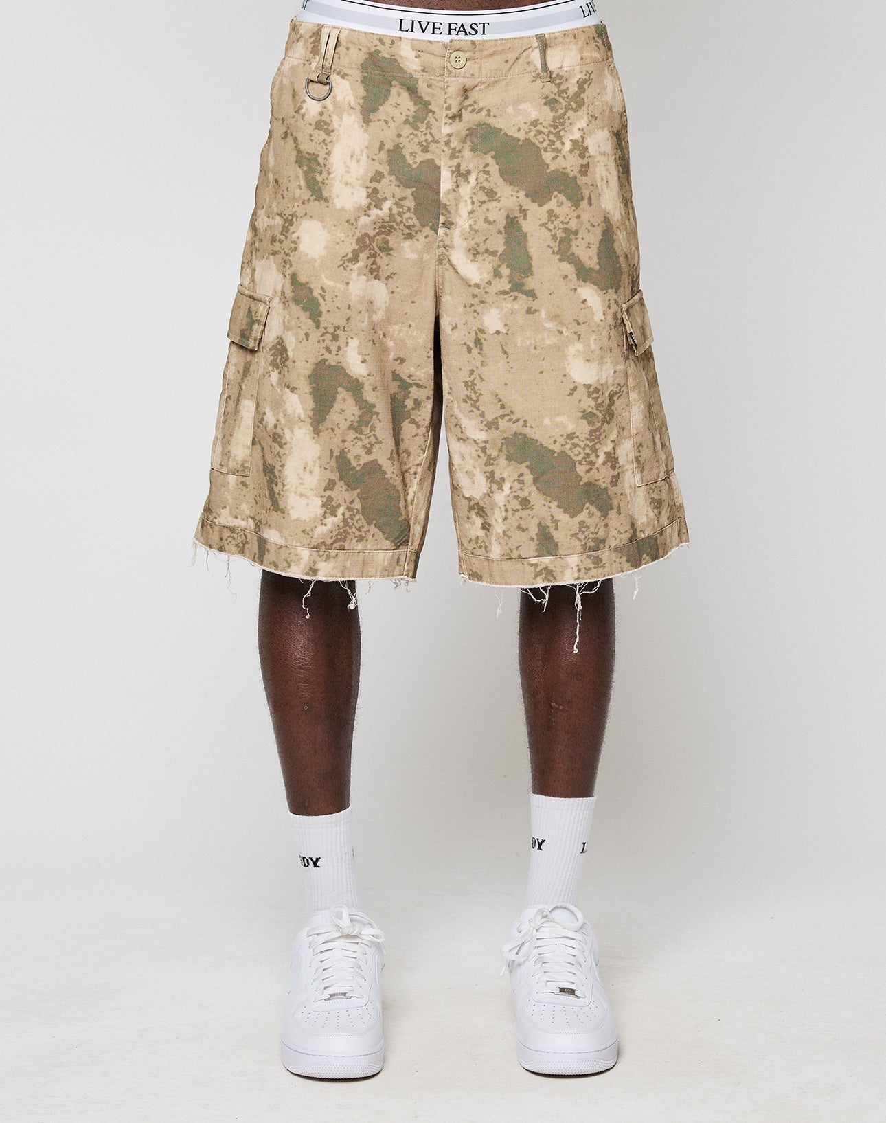 A person wears LFDY Cargo Shorts by LIVE FAST DIE YOUNG in tan and green camo with frayed hems, paired with a white LIVE FAST waistband, white socks, and white sneakers, showing a vintage style against a plain white background.
