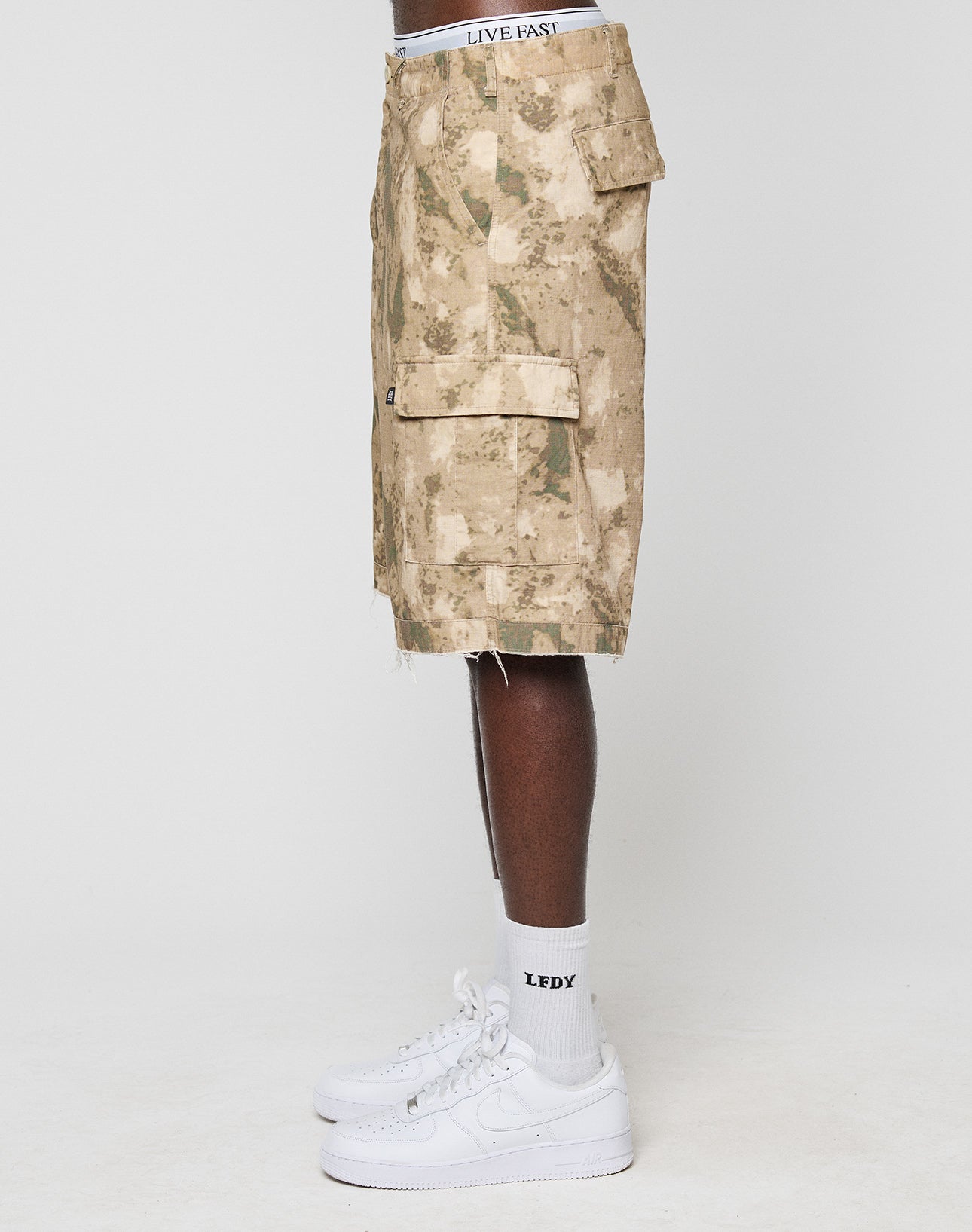 A person stands sideways wearing LFDY Cargo Shorts by LIVE FAST DIE YOUNG in beige camo with “LIVE FAST” on the 100% cotton waistband, paired with white “LEXX” socks and white sneakers against a plain white background.