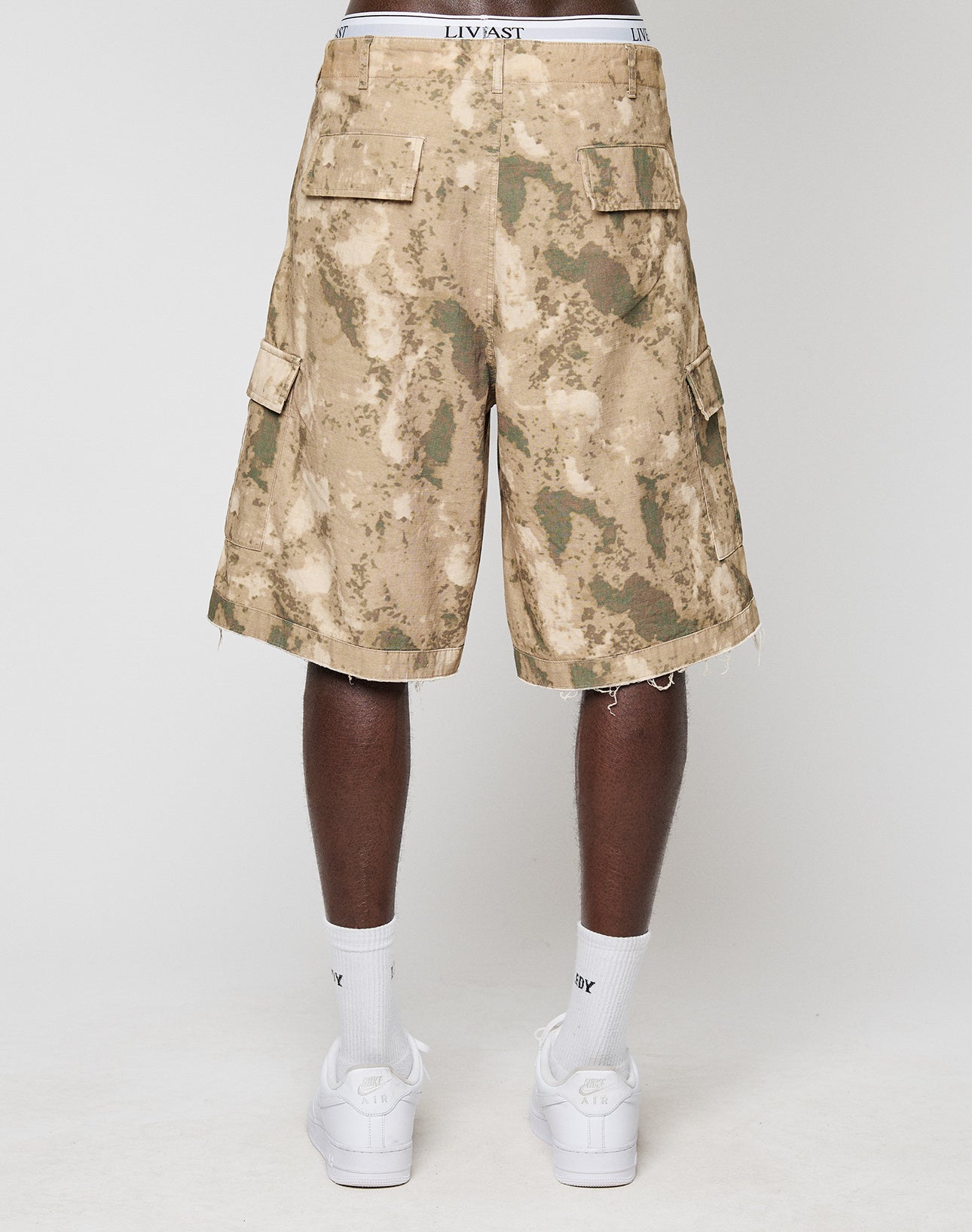 A person faces away wearing LFDY Cargo Shorts by LIVE FAST DIE YOUNG in vintage tie-dye tan and green 100% Baumwolle, a white LIVAST-branded waistband, white crew socks, and white sneakers against a plain light background.