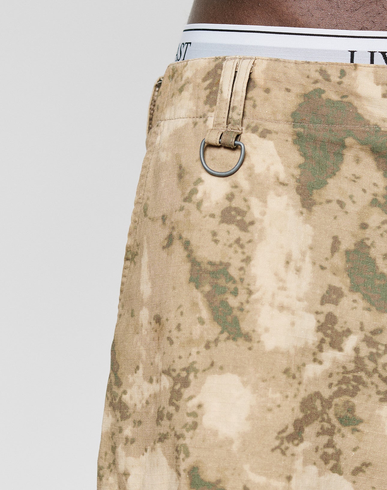 Close-up of a person in vintage style, wearing tan and green LFDY Cargo Shorts by LIVE FAST DIE YOUNG featuring a metal ring on the waistband; white underwear with black text peeks out above the shorts.