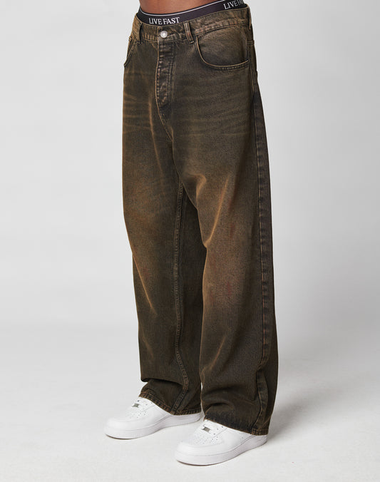 A person wearing LIVE FAST DIE YOUNGās LFDY Baggy Denimāloose, dark brown jeans with a faded wash and baggy fitāpaired with white sneakers and black underwear reading LIVE FAST, standing against a plain white background.