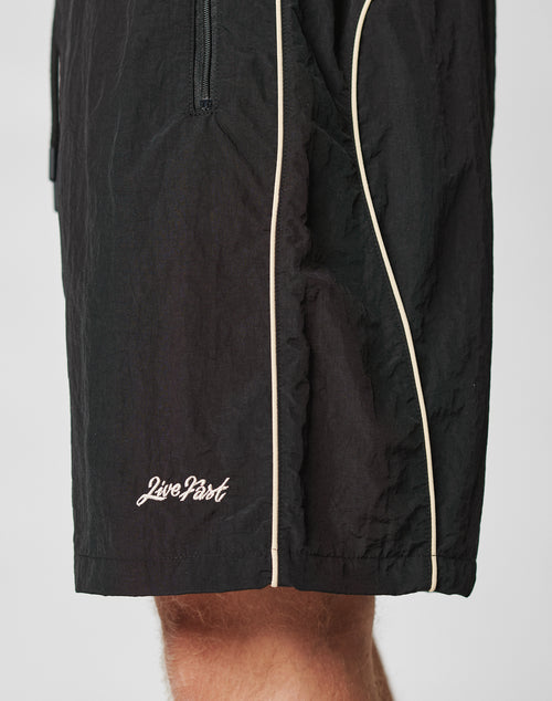 A close-up of a person wearing LIVE FAST DIE YOUNG Beach Shorts in black with white piping, featuring a mesh inlay and “Live Fast” embroidered in white near the hem.