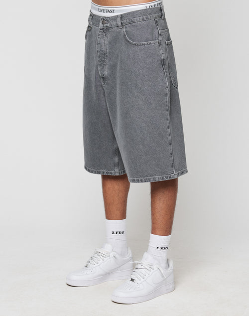 A person wearing LIVE FAST DIE YOUNG LFDY Jortsā100% Baumwolle gray denim shorts with a baggy fit and straight legāplus white crew socks with black text, white sneakers, and a visible white LIVE FAST waistband against a plain background.