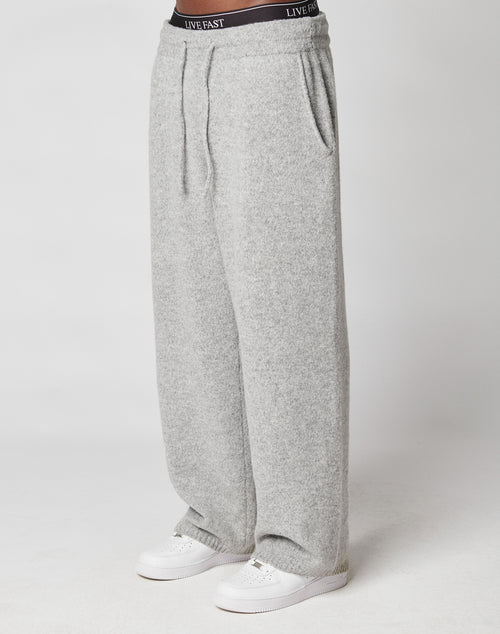 A person wearing LIVE FAST DIE YOUNG’s Live Fast Boucle Pants—loose, light gray polyester sweatpants with wide fit, elastic waistband reading LIVE FAST—hands in pockets and white sneakers, poses against a plain white background.