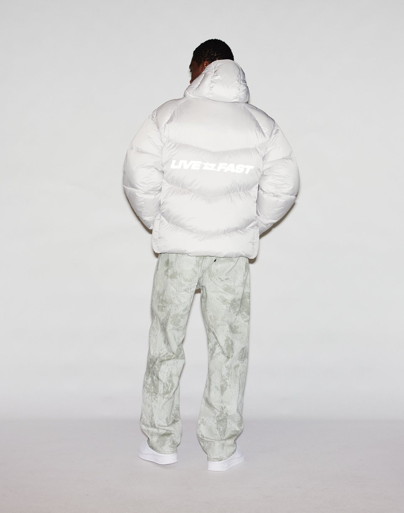 A person faces away wearing the Live Fast Puffer Jacket by LIVE FAST DIE YOUNG, featuring a bold LIVE FAST reflektierender Print on the back, paired with marbled pants and white shoes against a light background.