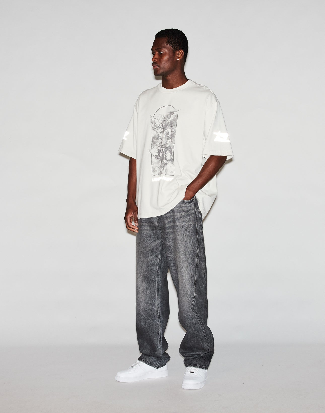 A man stands against a white background wearing the LFDY Window Tee by LIVE FAST DIE YOUNG, featuring reflective sleeve and logo prints, paired with baggy gray jeans and white sneakers. He looks slightly to the side with his hands in his pockets.