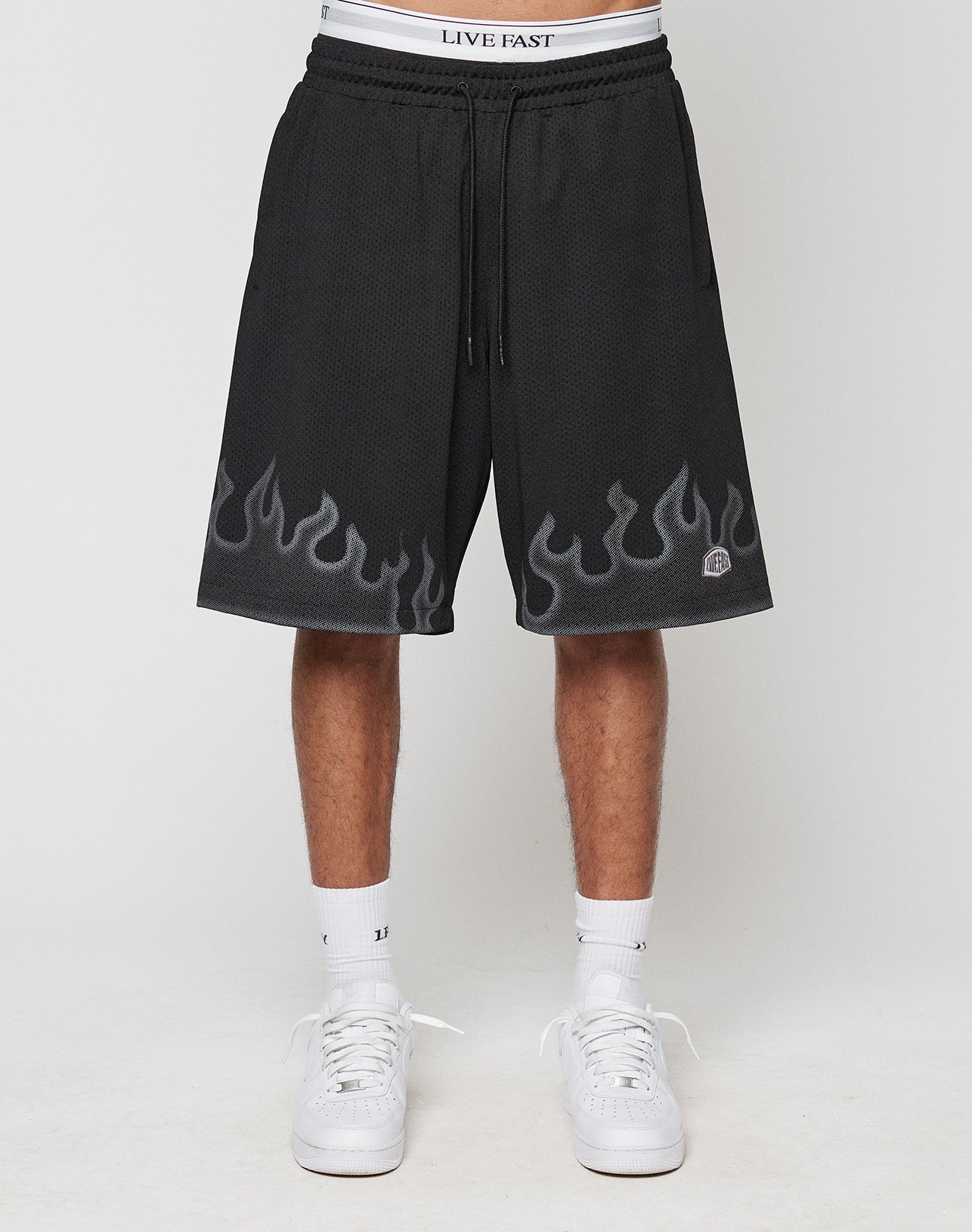 A person wears LIVE FAST DIE YOUNG Burning Mesh Shorts with gray flame print along the hem, LIVE FAST on the elastic waistband, paired with white socks and sneakers. Photo shows a plain background.