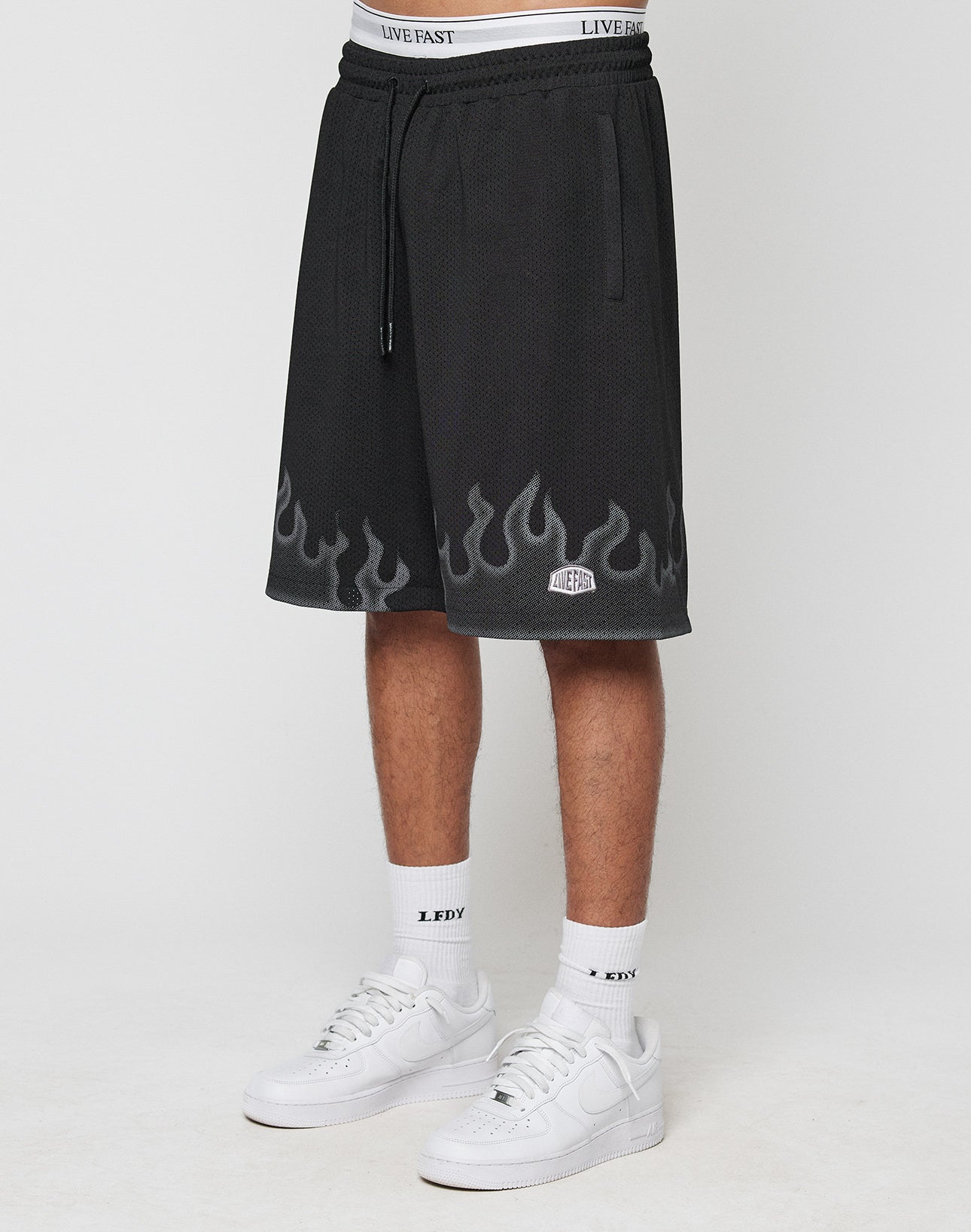 A person wears LIVE FAST DIE YOUNG Burning Mesh Shorts with a Flammenprint hem and elastic waistband, showing the white underwears band above. Paired with white socks and sneakers, theyre photographed against a plain background.