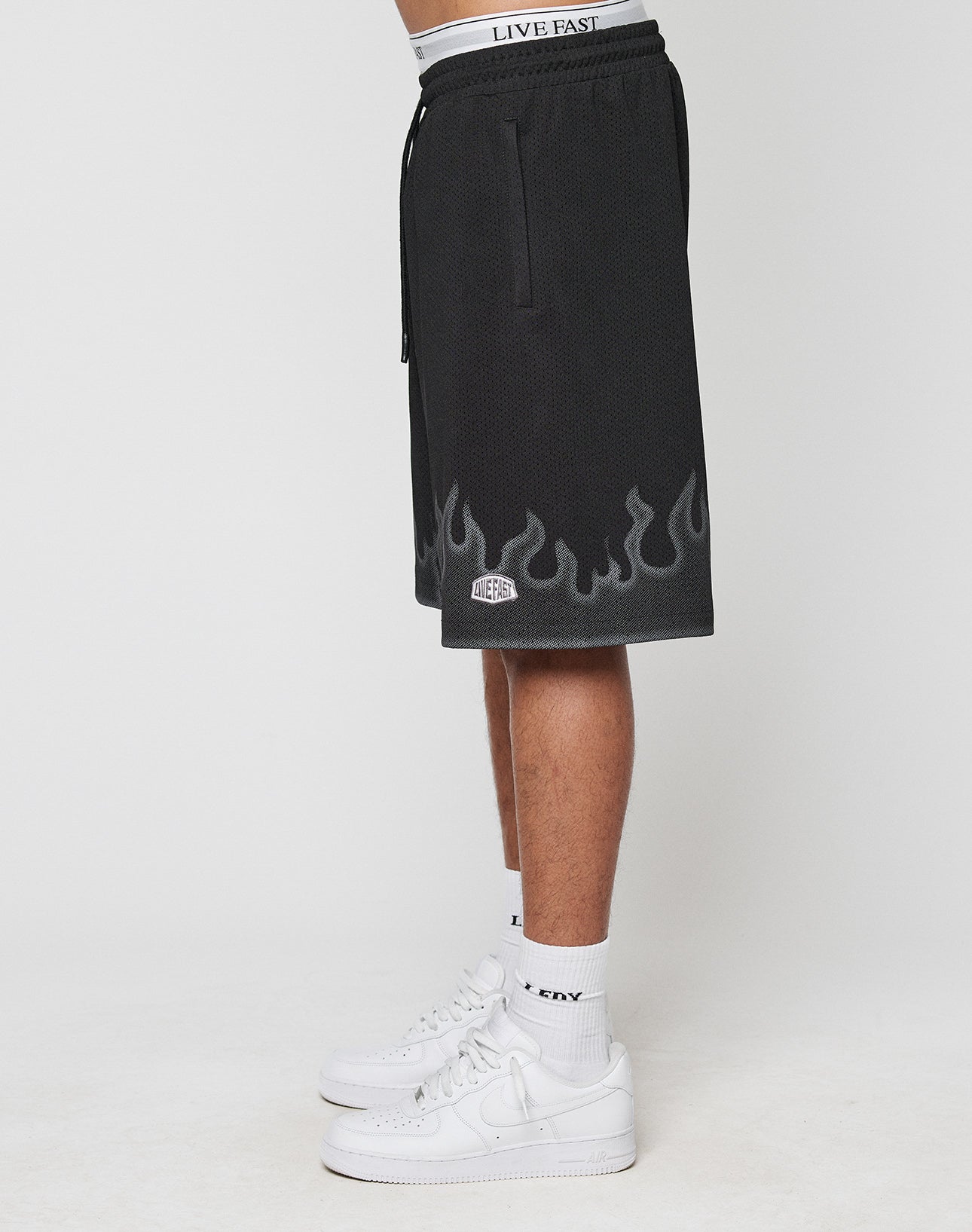 A person stands in profile against a white background wearing LIVE FAST DIE YOUNG Burning Mesh Shorts with Flammenprint and an elastic waistband reading LIVE FAST, paired with white crew socks and white Nike sneakers.