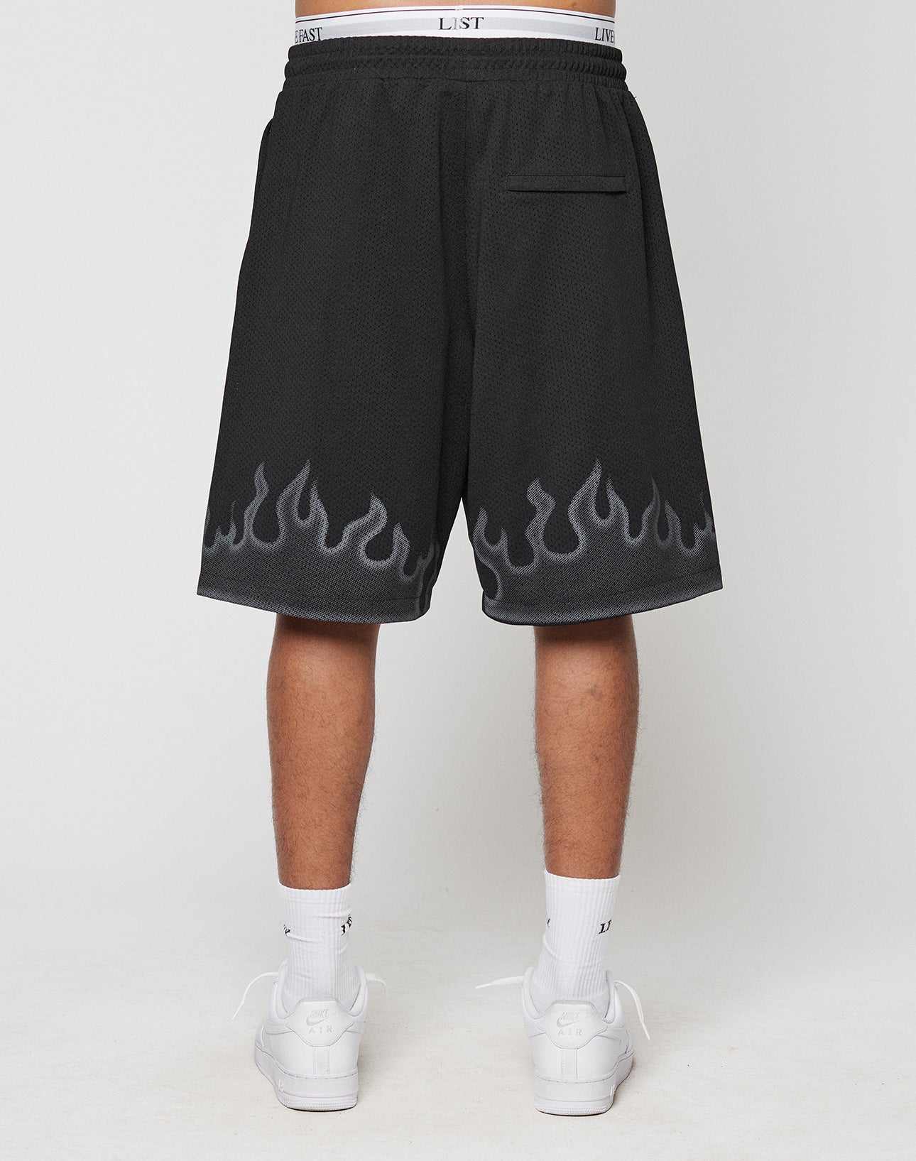A person stands facing away in LIVE FAST DIE YOUNG Burning Mesh Shorts with an Oversize Fit and gray Flammenprint at the hem, paired with white socks and sneakers, against a plain white background.