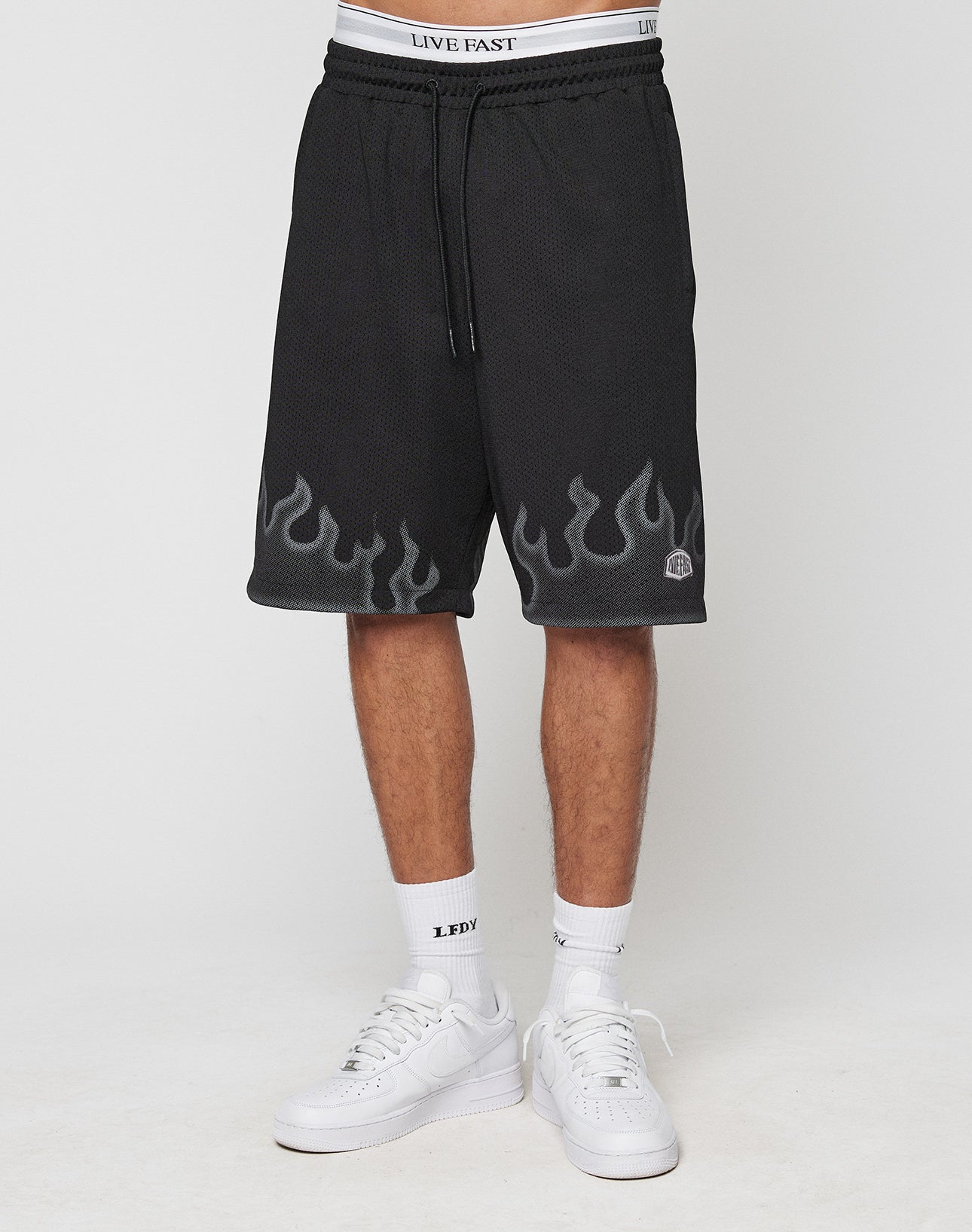 A person wears LIVE FAST DIE YOUNG Burning Mesh Shorts with a gray Flammenprint and LIVE FAST waistband, white LEDY socks, and white sneakers, standing against a plain white background.
