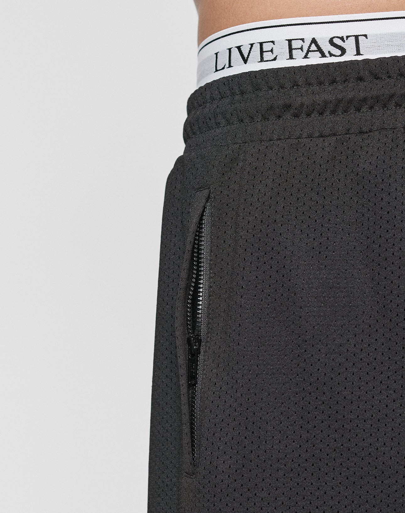 Close-up of a person wearing Burning Mesh Shorts by LIVE FAST DIE YOUNG, featuring an elastic waistband and zippered pocket. The white underwear waistband with LIVE FAST is visible above the shorts.