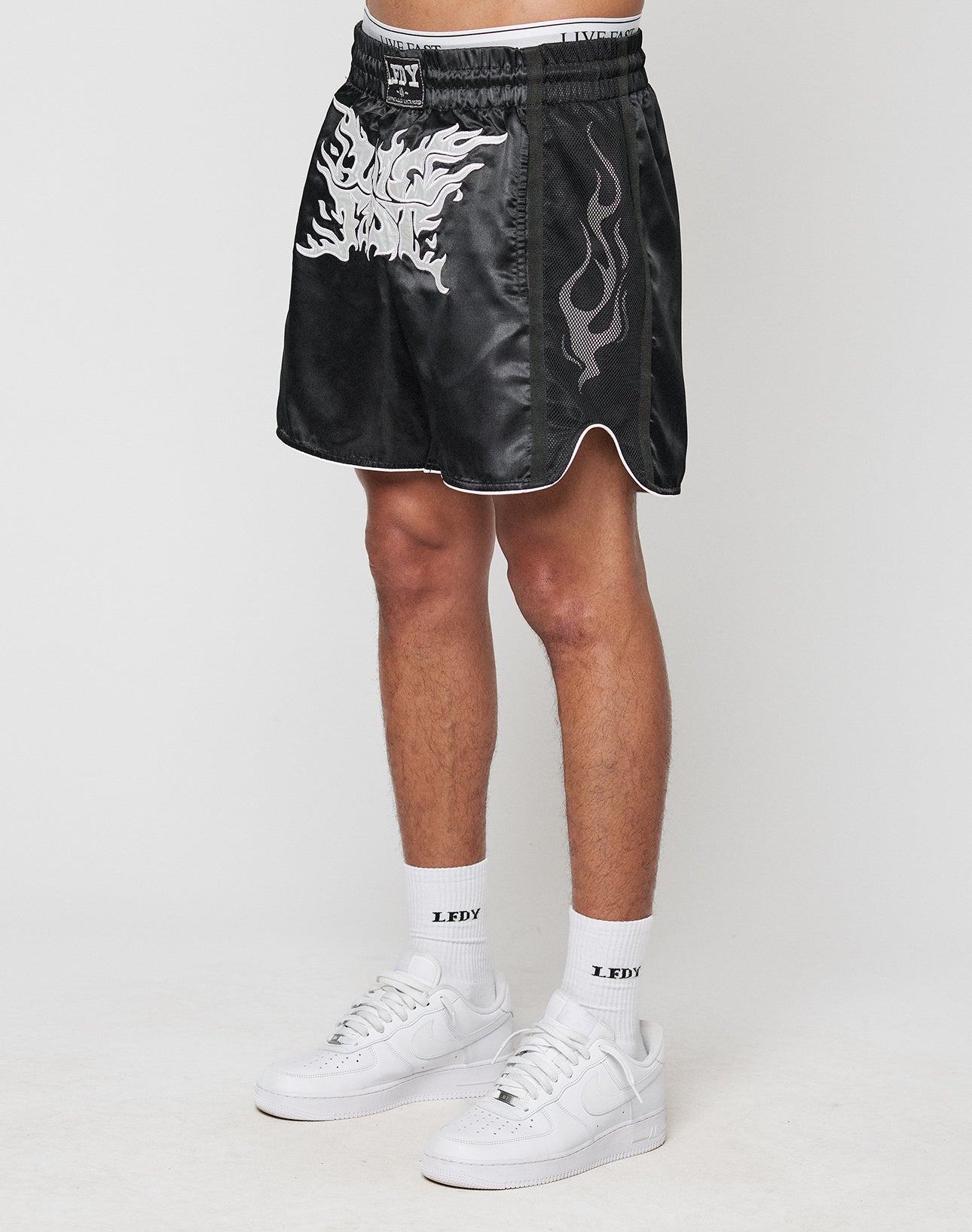 A person wears LIVE FAST DIE YOUNG LFDY Fighting Shorts—black with white flames and an elastic waistband—paired with white LFDY socks and white sneakers. Only their lower half is shown against a plain white background.