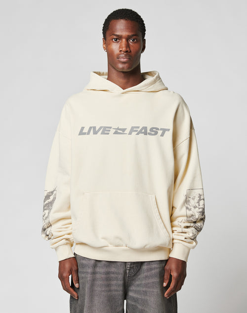 A man wears the LFDY Window Hooded by LIVE FAST DIE YOUNGāan oversized, cream hoodie with bold gray LIVE FAST lettering and graphic sleeve designs, featuring reflektierender Druck. He stands against a plain light background.