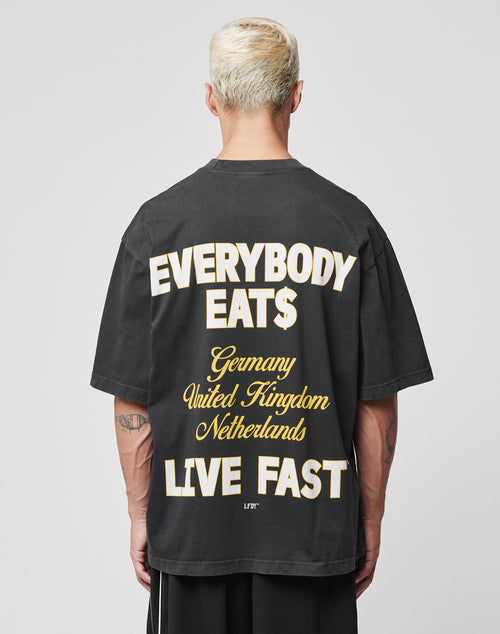A person with short, bleached blond hair wears the LIVE FAST DIE YOUNG Everybody Eats Tee, a black Oversize Fit shirt made of 100% Baumwolle with bold yellow and white text on the back. Plain gray background; care label includes waschhinweise.