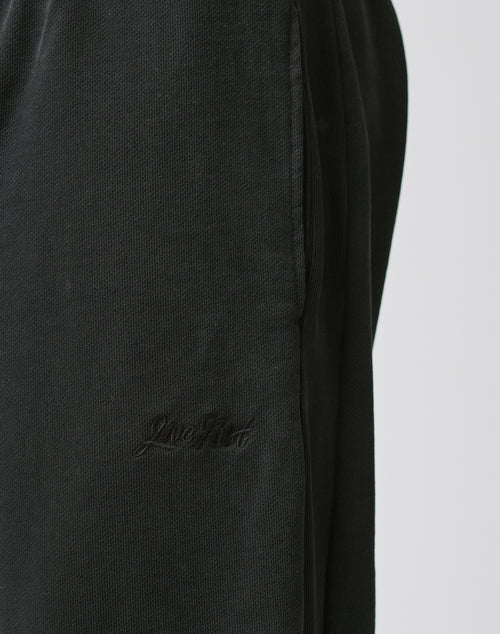 Close-up of Basic Wide Jogger by LIVE FAST DIE YOUNG in black textured cotton, with a wide fit, elastic waistband, and embroidered Live Good logo near the pocket. Shown against a plain, light-colored background.