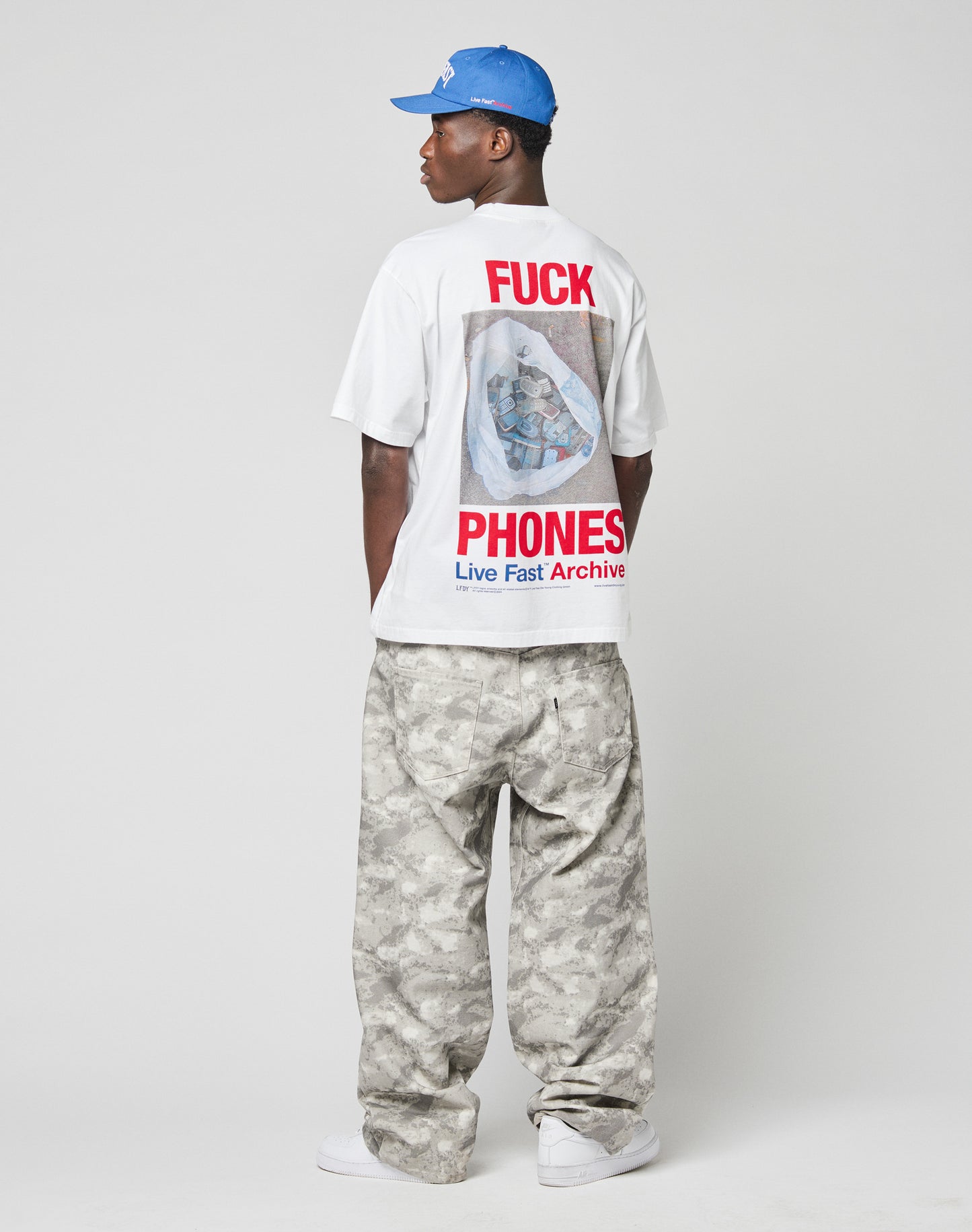 A man faces away, wearing the LIVE FAST DIE YOUNG LFDY F*ck Phones Tee with front and back prints, a blue cap, baggy gray camo pants, and white sneakers against a plain light background.
