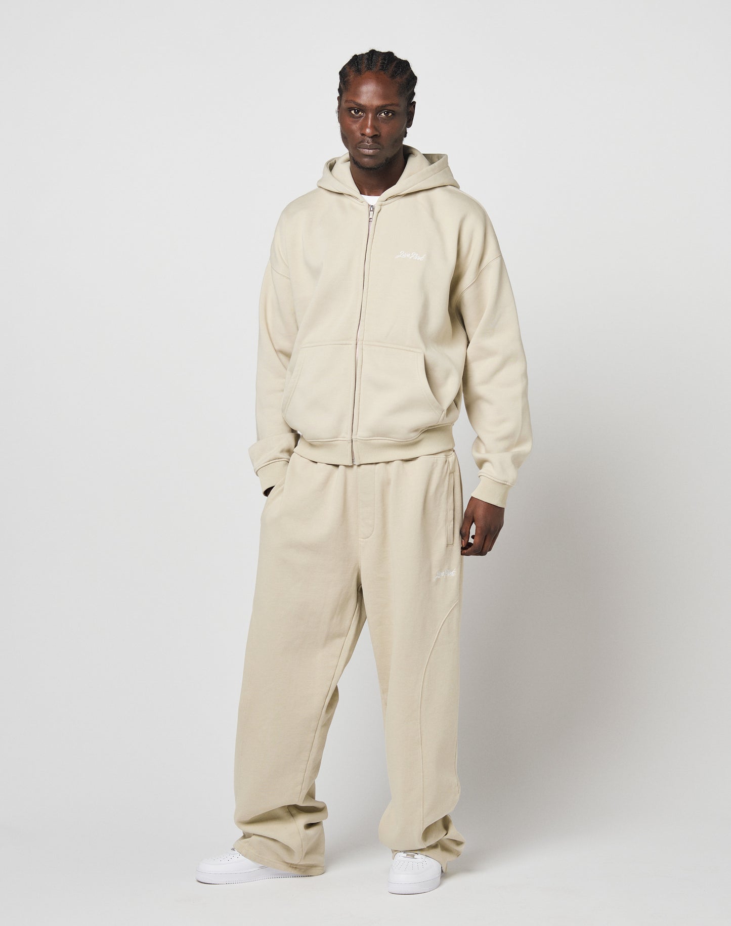 A person poses against a white background, wearing the LIVE FAST DIE YOUNG Basic Script Hooded Zip sweatshirt in beige with matching sweatpants and white sneakers—an oversized, relaxed look ideal for everyday comfort.