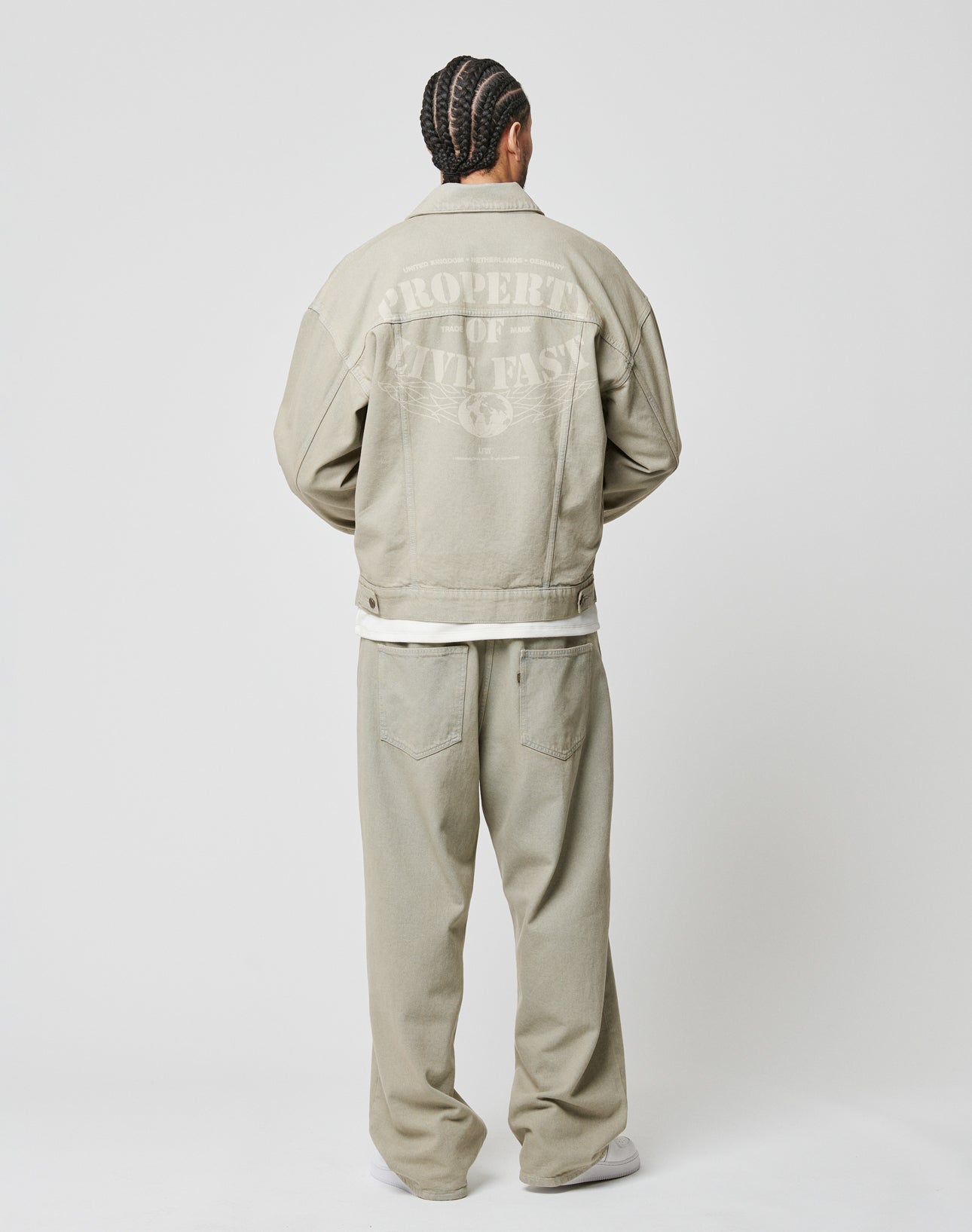 A person with braided hair faces away, wearing the LIVE FAST DIE YOUNG Live Fast Denim Jacket in light beige, 100% cotton, with an oversized fit and laser-printed PROPERTY OF LIVE FAST graphic on the back. Background is plain white.