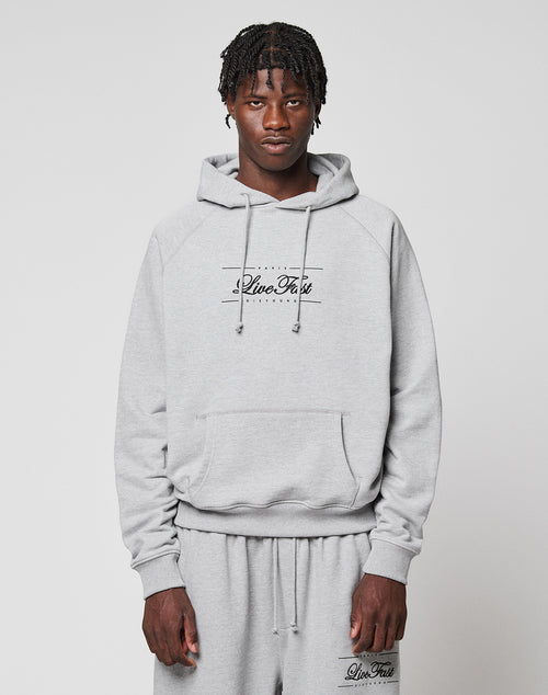 A young man with short braids wears a light gray LFDY Embroidery Hooded by LIVE FAST DIE YOUNG, paired with matching sweatpants. Both pieces feature "Live Fast" and he stands against a plain white background, looking at the camera.