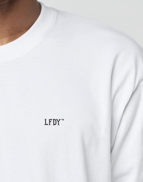 A close-up of a person in the LIVE FAST DIE YOUNG Basic Chest Tee, a plain white crew-neck shirt made from 100% cotton with an embroidered black LFDY™ logo on the chest. Only their lower face and upper torso are visible.