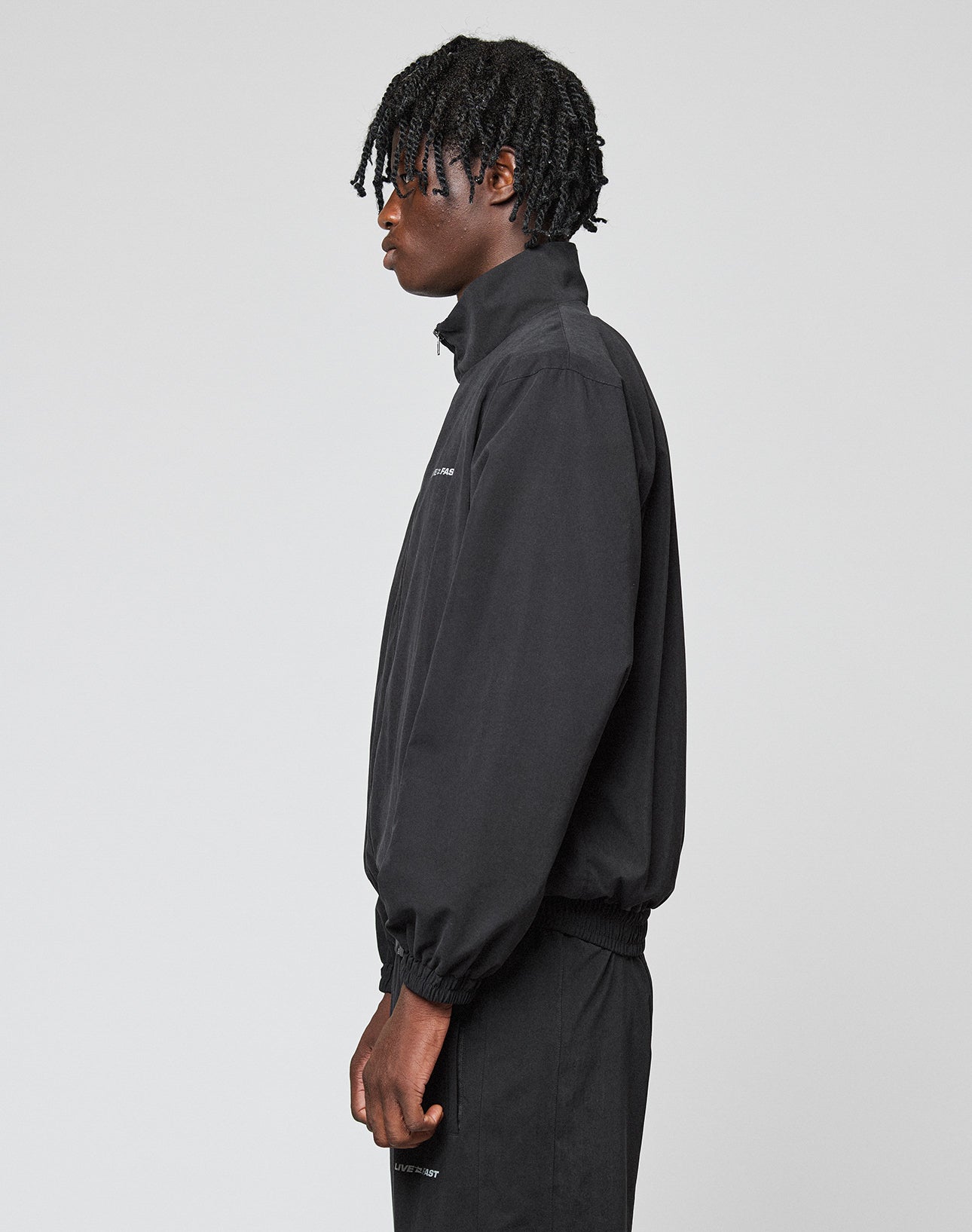 A person with short, twisted black hair stands in profile against a light grey background, wearing an oversize fit black LFDY Trackjacket by LIVE FAST DIE YOUNG with subtle Logo-Stick details and matching black pants.