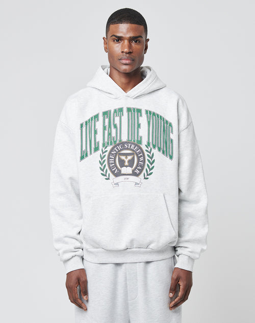 A man stands against a light gray background, wearing the LIVE FAST DIE YOUNG Hooded by LIVE FAST DIE YOUNG—an oversize fit hoodie in light gray, featuring a green front print and crest, made from a soft Baumwolle-Polyester mix.
