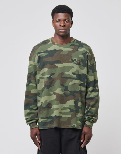 A man stands facing forward in LIVE FAST DIE YOUNGs Copyright Waffle Longsleeve, an oversize fit camo shirt with bold back print, paired with black pants, set against a plain light gray background.