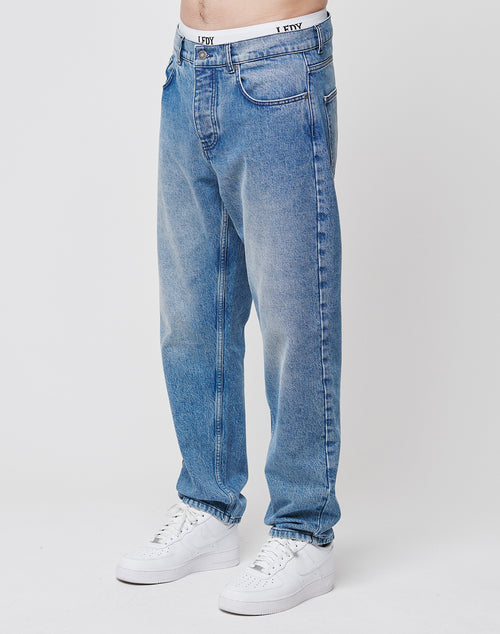 A person stands against a plain white background wearing LIVE FAST DIE YOUNG’s Regular Denim jeans in light blue, paired with white sneakers and a white waistband with black text; the image is cropped just above the waist.