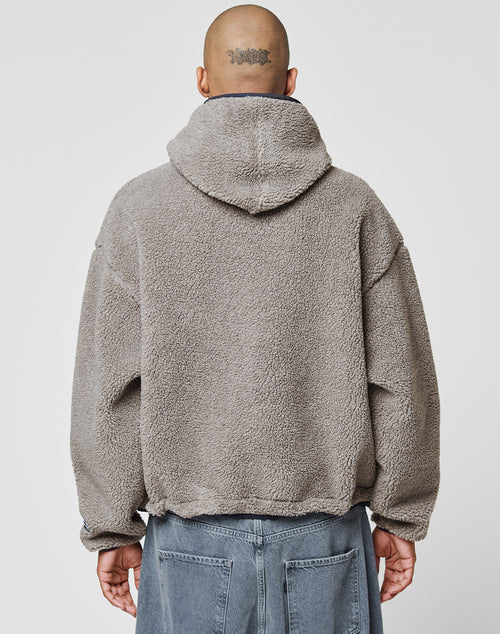 A person with a shaved head and a tattoo wears the LIVE FAST DIE YOUNG LFDY Teddy Hooded jacket, featuring an oversized fit and Eingrifftaschen, paired with denim pants, standing with their back to a plain white background.