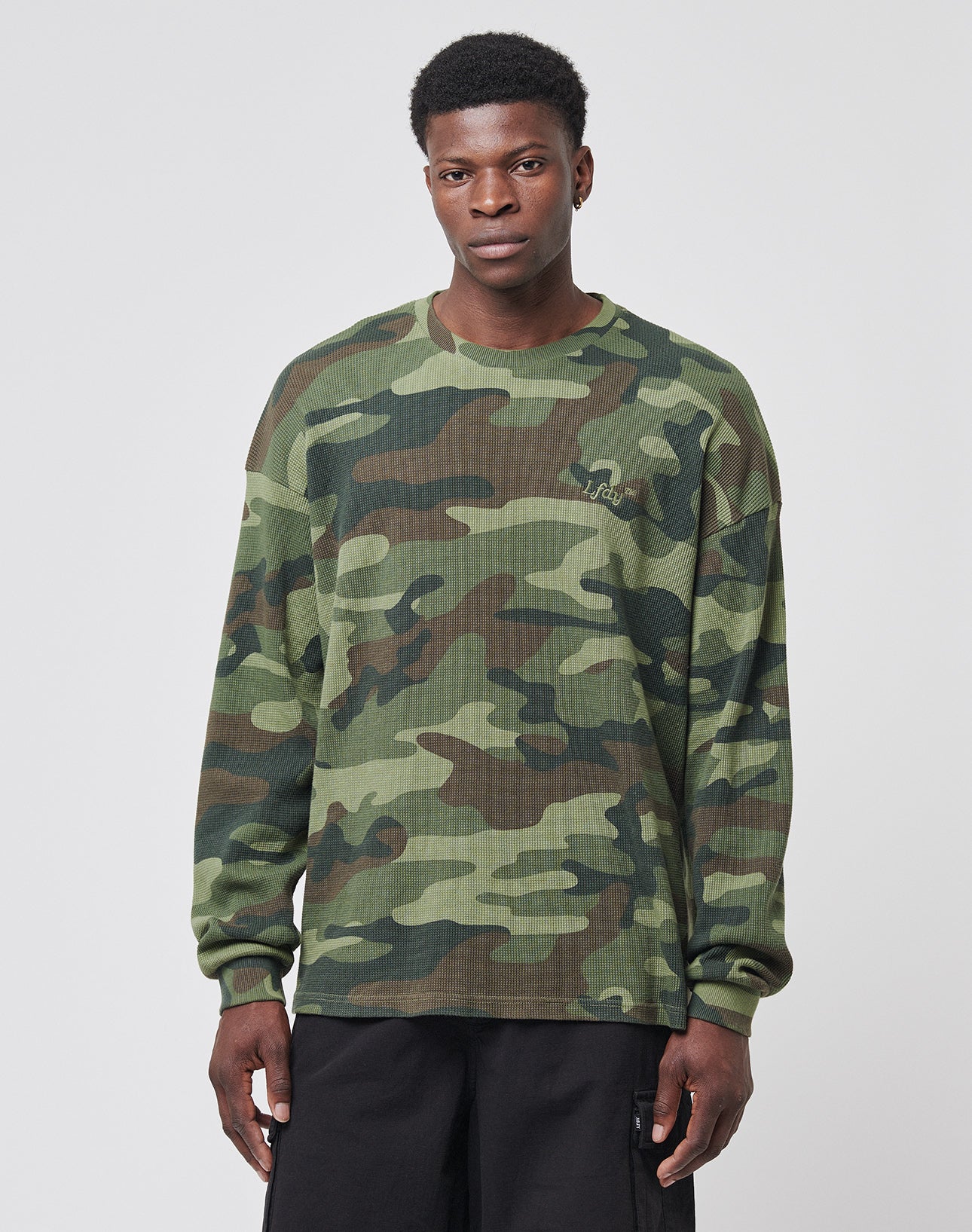 A man stands against a plain background, wearing the LIVE FAST DIE YOUNG Copyright Waffle Longsleeve in an oversize green and brown camouflage pattern with black pants, looking directly at the camera with a neutral expression.