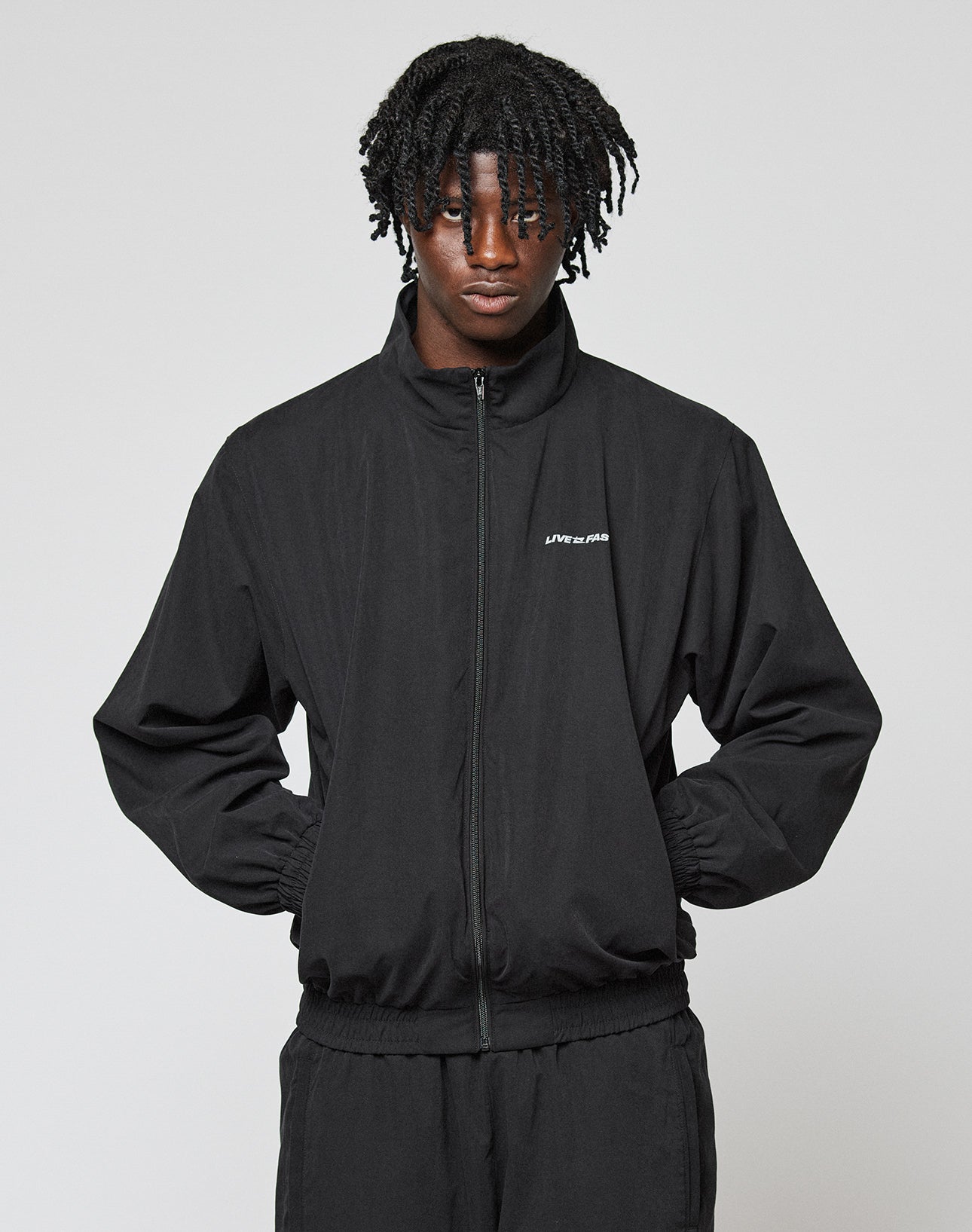 A person with medium-length, curly hair wears a black, oversize fit LFDY Trackjacket and matching pants from LIVE FAST DIE YOUNG. The jacket features LIVESTRONG text on the chest. They stand with hands in pockets against a plain background.