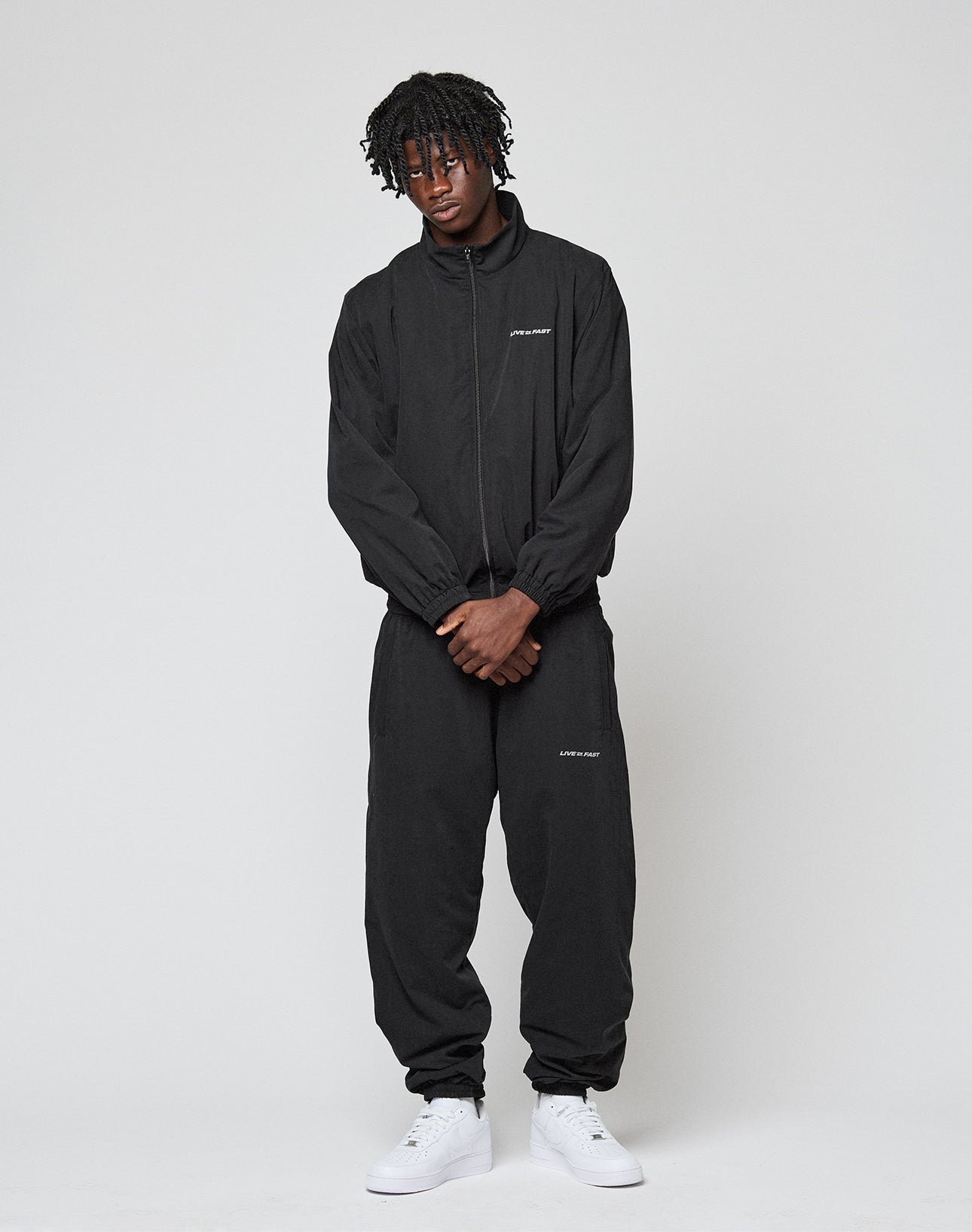 A person with short dreadlocks stands against a plain background, wearing the black LFDY Trackjacket by LIVE FAST DIE YOUNG in an oversize fit with white logo text, paired with white sneakers and looking relaxed with hands together at the waist.