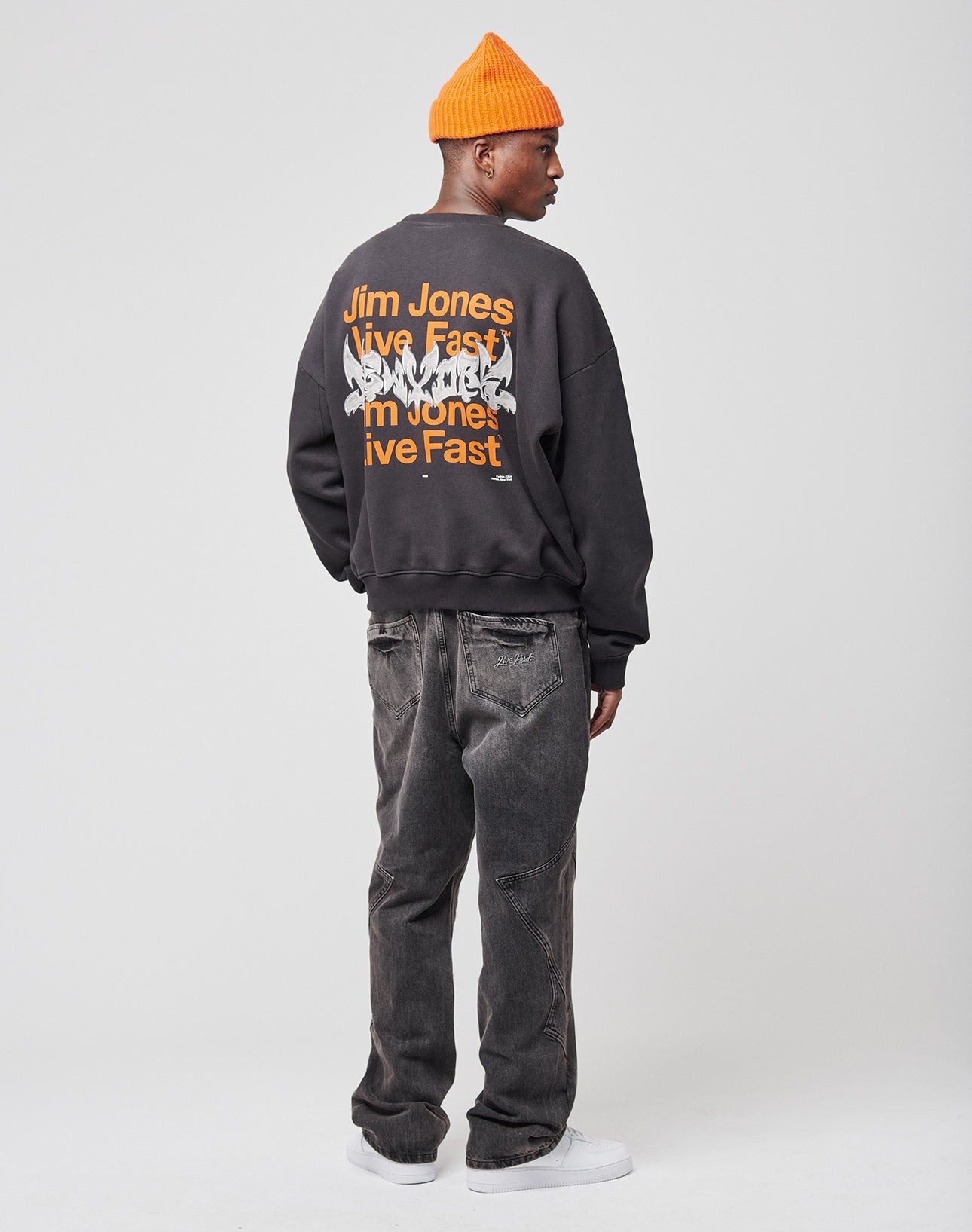 A person wears the LIVE FAST DIE YOUNG Live Fast NY Sweater in an oversized fit with bold front and back “Jim Jones Live Fast” prints, paired with a bright orange beanie, black jeans, and white sneakers against a plain white background.