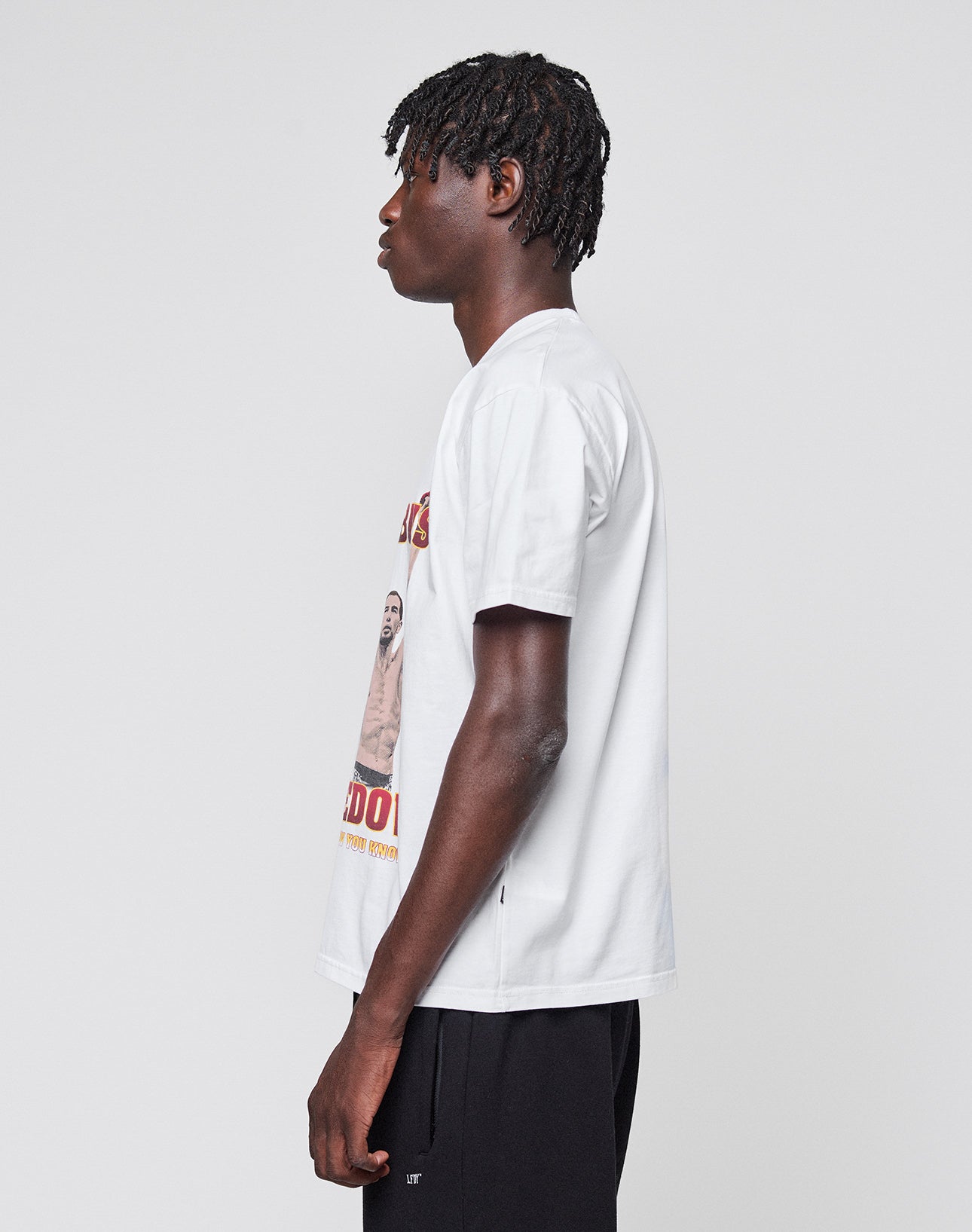 A young man with short dreadlocks wears the Abus Magomedov Tee by LIVE FAST DIE YOUNG, a white T-shirt with front and back print in 100% Baumwolle, paired with black shorts, standing in profile against a plain light gray background.
