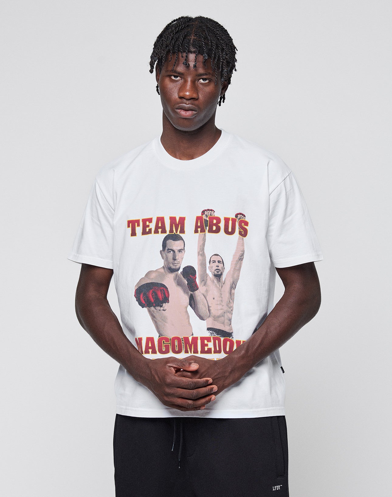 A man with short, twisted hair stands against a plain backdrop, wearing the LIVE FAST DIE YOUNG Abus Magomedov Tee, a white 100% cotton Regular Fit shirt featuring two boxers and "TEAM ABUS NAGOMEDOV" in red and yellow letters.