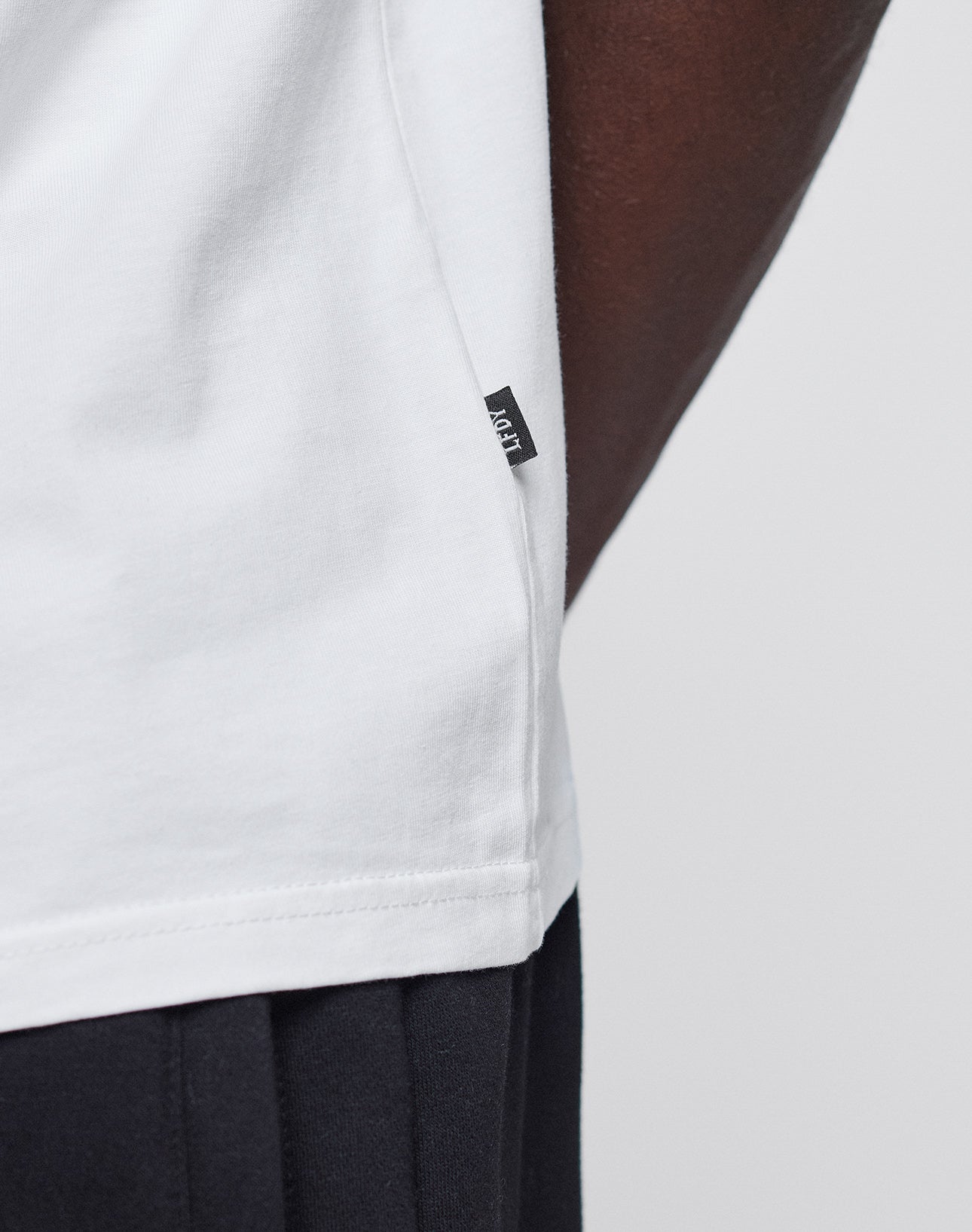Close-up of a person wearing the Abus Magomedov Tee by LIVE FAST DIE YOUNG, a white 100% Baumwolle Regular Fit T-shirt with a small black fabric tag on the side seam, paired with dark pants against a plain, light background.