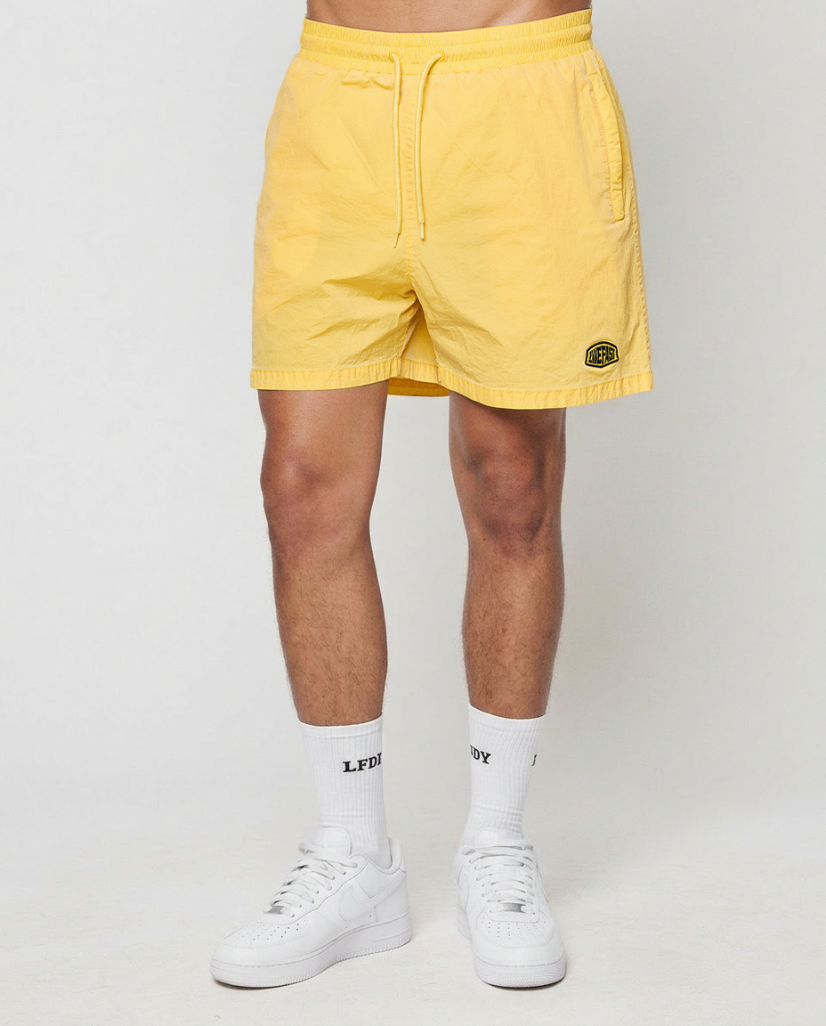 A person in LIVE FAST DIE YOUNG's Live Fast Swim Shorts (light yellow, regular fit, elastic waistband and drawstring) pairs them with white socks and sneakers, standing against a plain background.
