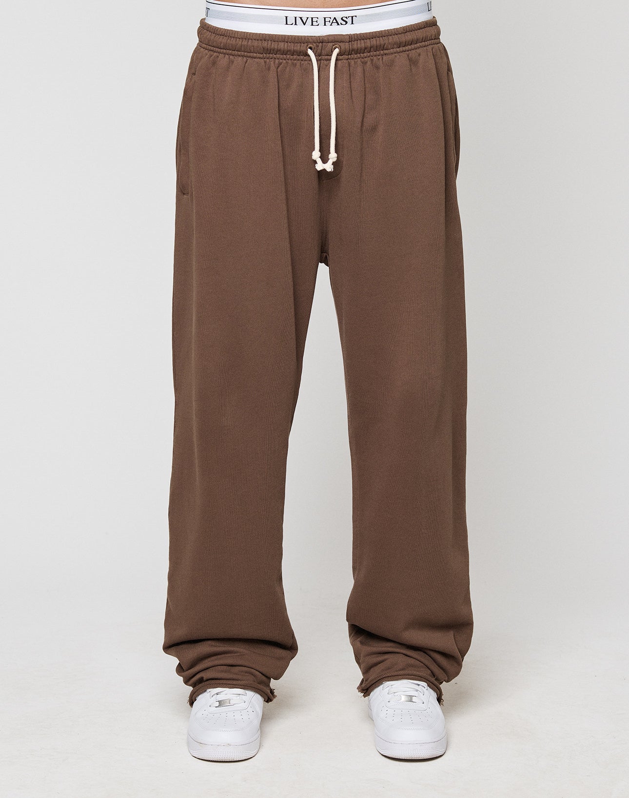 A person wears LFDY Lightweight Jogger by LIVE FAST DIE YOUNG—loose brown 100% cotton sweatpants with a white drawstring, white sneakers, and a LIVE FAST waistband, set against a plain, light background.