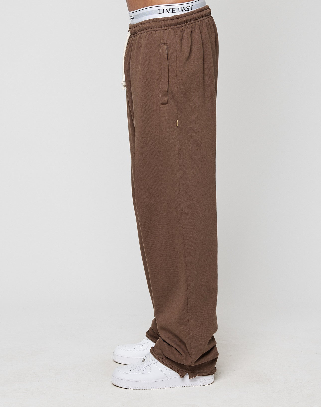 A person wearing loose-fitting LFDY Lightweight Jogger sweatpants by LIVE FAST DIE YOUNG, white sneakers, and a visible white waistband with LIVE FAST text, standing sideways against a plain white background.