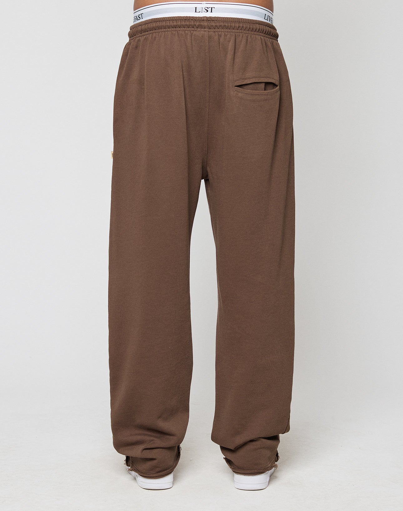 A person wears LIVE FAST DIE YOUNG’s LFDY Lightweight Jogger—loose-fitting brown French Terry sweatpants with an elastic waistband, visible white underwear, and a small back pocket—standing on a light background.